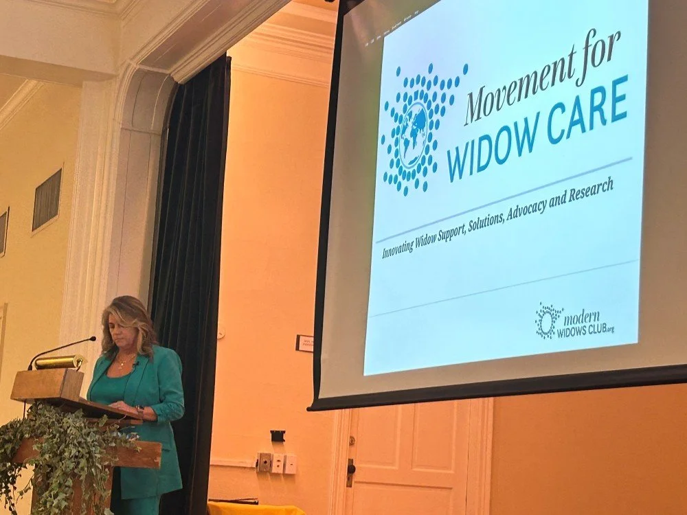 A woman giving a presentation at a conference, standing behind a wooden podium with a greenery decoration, next to a large screen displaying a slide about 'Movement for Widow Care' with the subtitle 'Innovating Widow Support, Solutions, Advocacy and Research.'