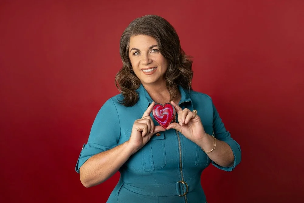 A woman with shoulder-length brown hair, wearing a teal button-up dress, smiling and holding a heart-shaped object with a red and pink pattern over her chest, against a red background.
