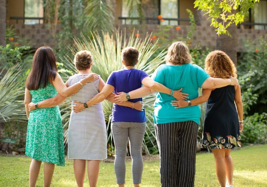 Group of six women standing outdoors with arms around each other's shoulders and waists, facing away from the camera, in a lush garden.