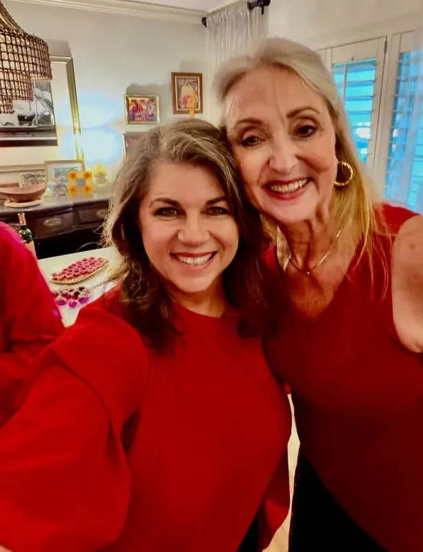 Carolyn and another woman smiling and taking a selfie in a cozy, decorated room. Both are wearing red tops and appear joyful.