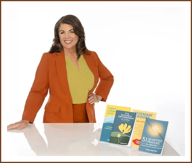 A woman with brown, wavy hair, wearing an orange blazer over a yellow blouse, smiling and standing behind a white table displaying several books about widow empowerment and spirituality.