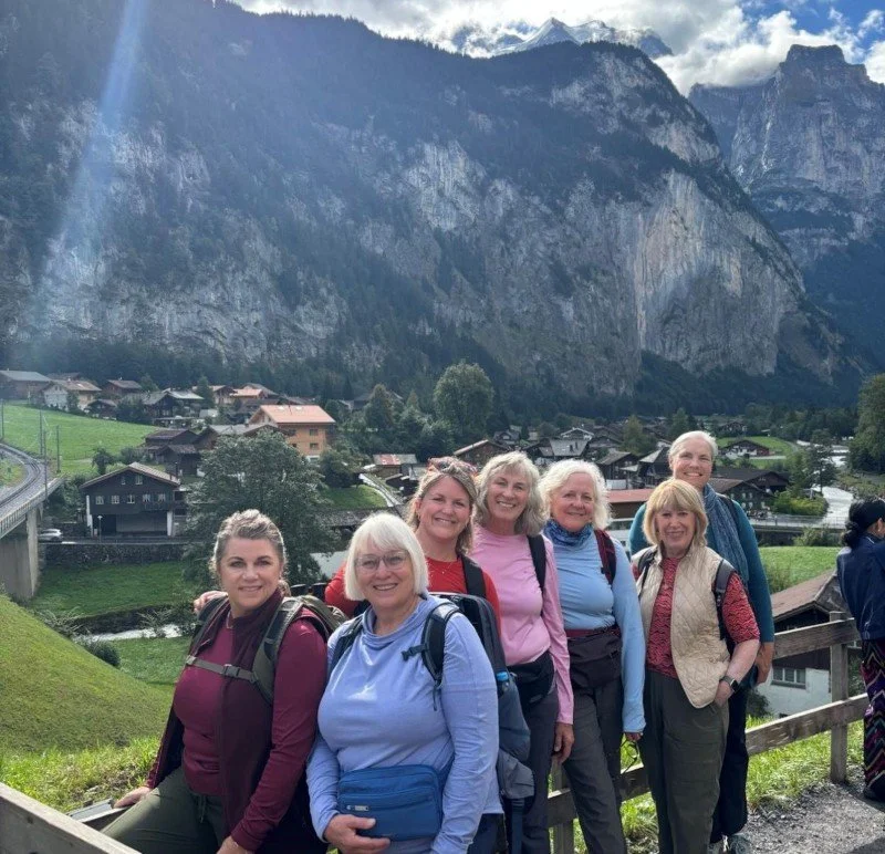 Carolyn and a group of six other women posing outdoors in a scenic mountainous area with houses, green fields, and cliffs in the background.
