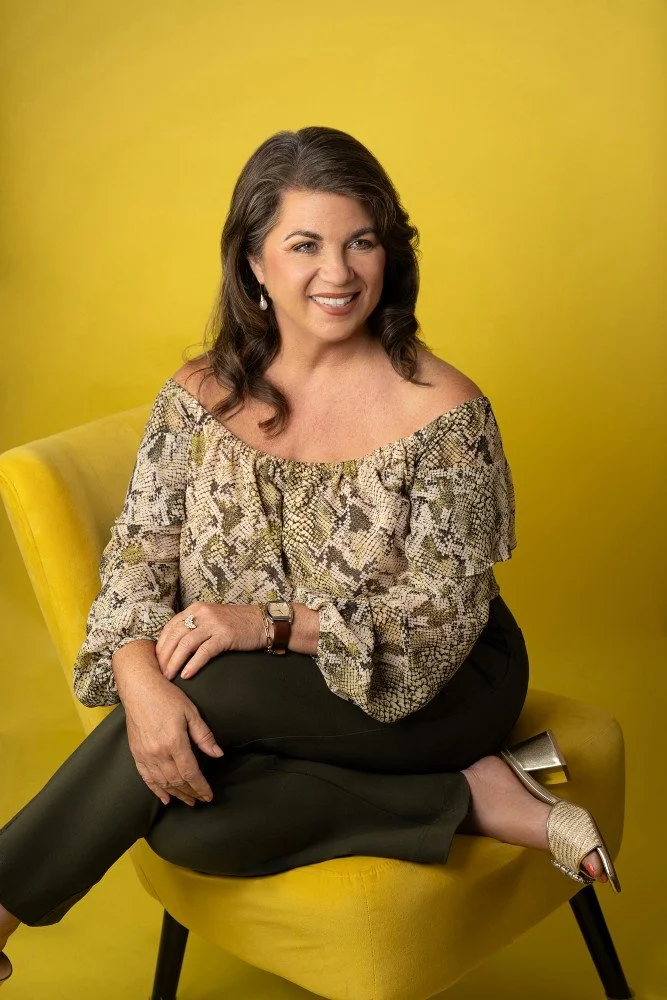 A woman with dark brown, wavy hair sitting on a yellow chair against a yellow background. She is smiling, wearing a snake print off-shoulder blouse, black pants, gold open-toe heels, and jewelry including earrings and a wristwatch.