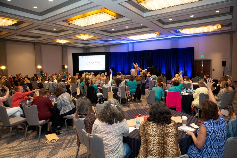 Large conference room filled with seated attendees, many with raised hands, facing a stage with a performer and large screen displaying 'Carolyn Moore.'