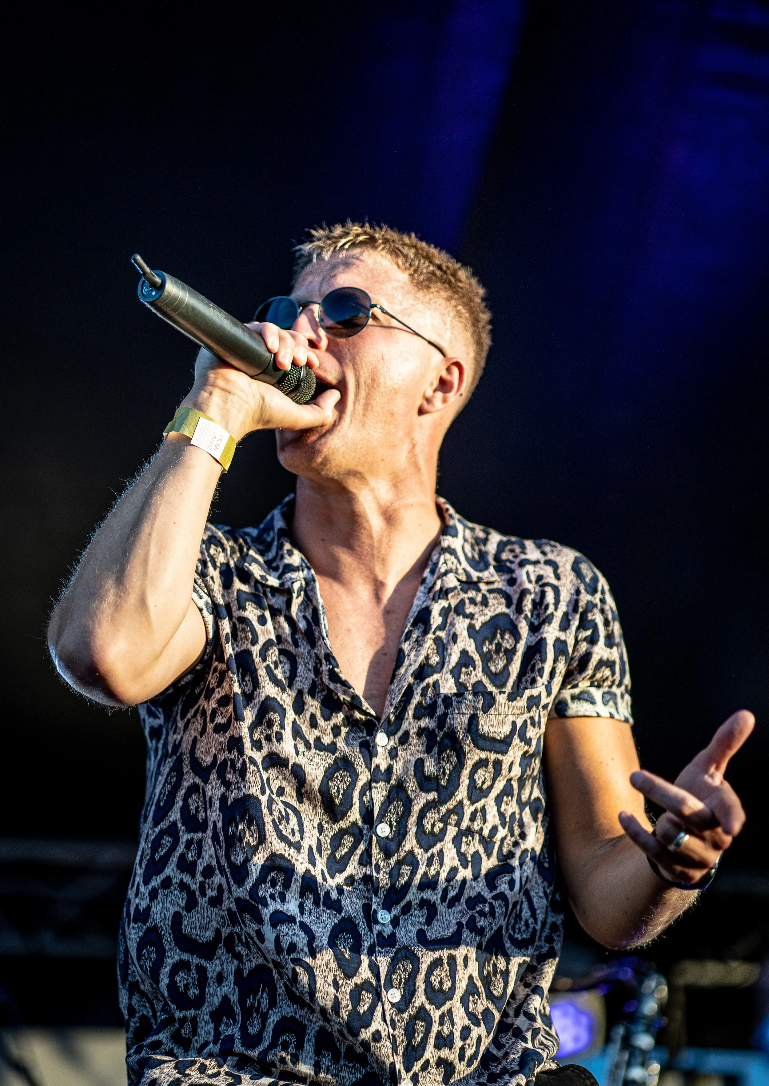 A man with short blonde hair wearing dark sunglasses and a leopard print shirt is holding a microphone and singing or speaking on stage with a dark background and stage lighting.