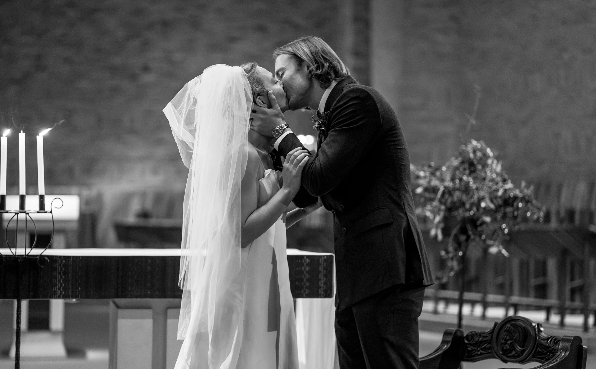 A black and white photograph of a couple kissing during a wedding ceremony. The bride is wearing a veil and a white dress, while the groom is in a dark suit. They are holding each other tenderly, with a background that includes lit candles and floral