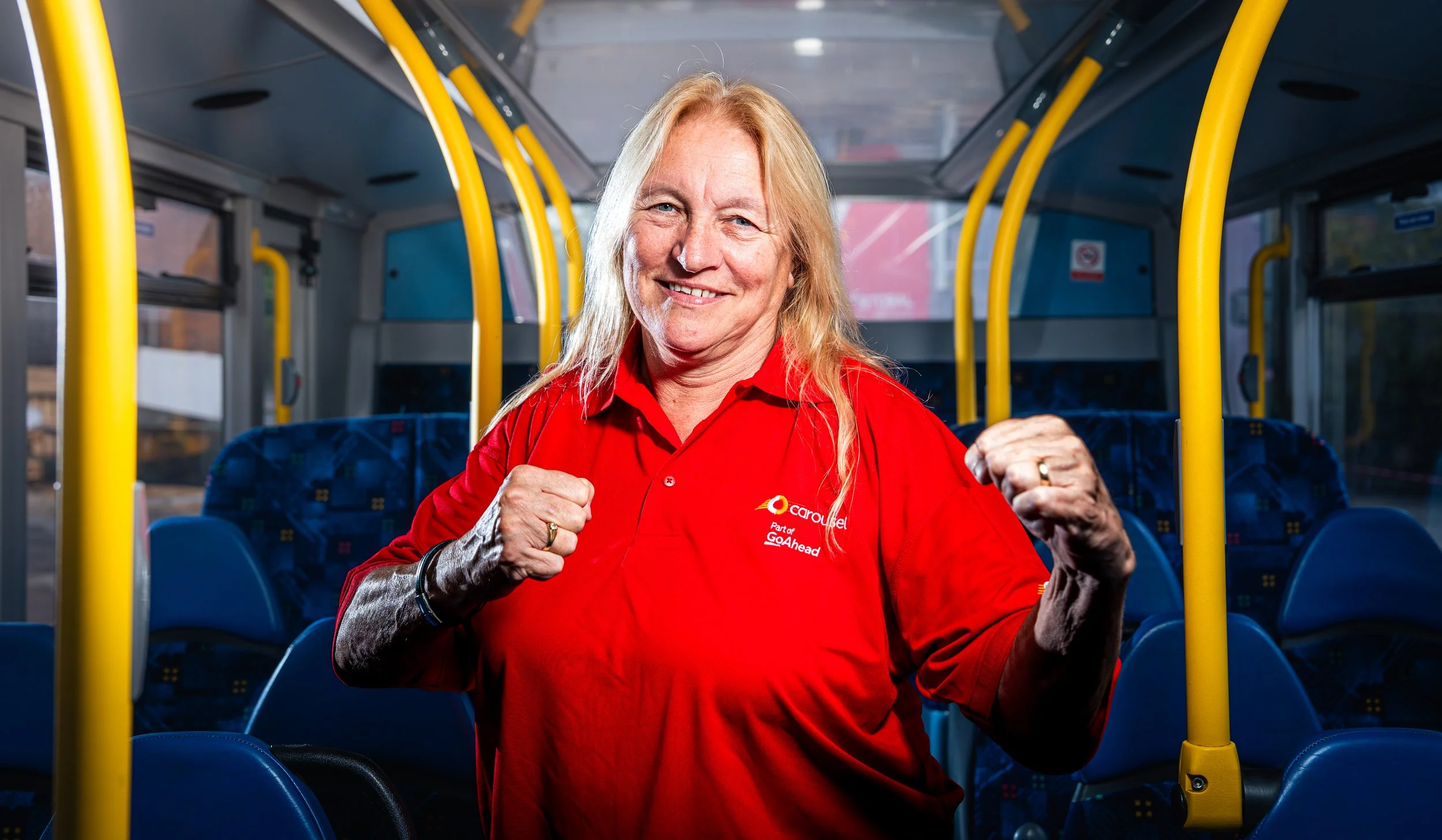 A smiling woman with long blonde hair, wearing a red uniform shirt with a Carousel Part of GoAhead logo, standing inside a bus with blue seats and yellow handrails, making a fists-clenching gesture.