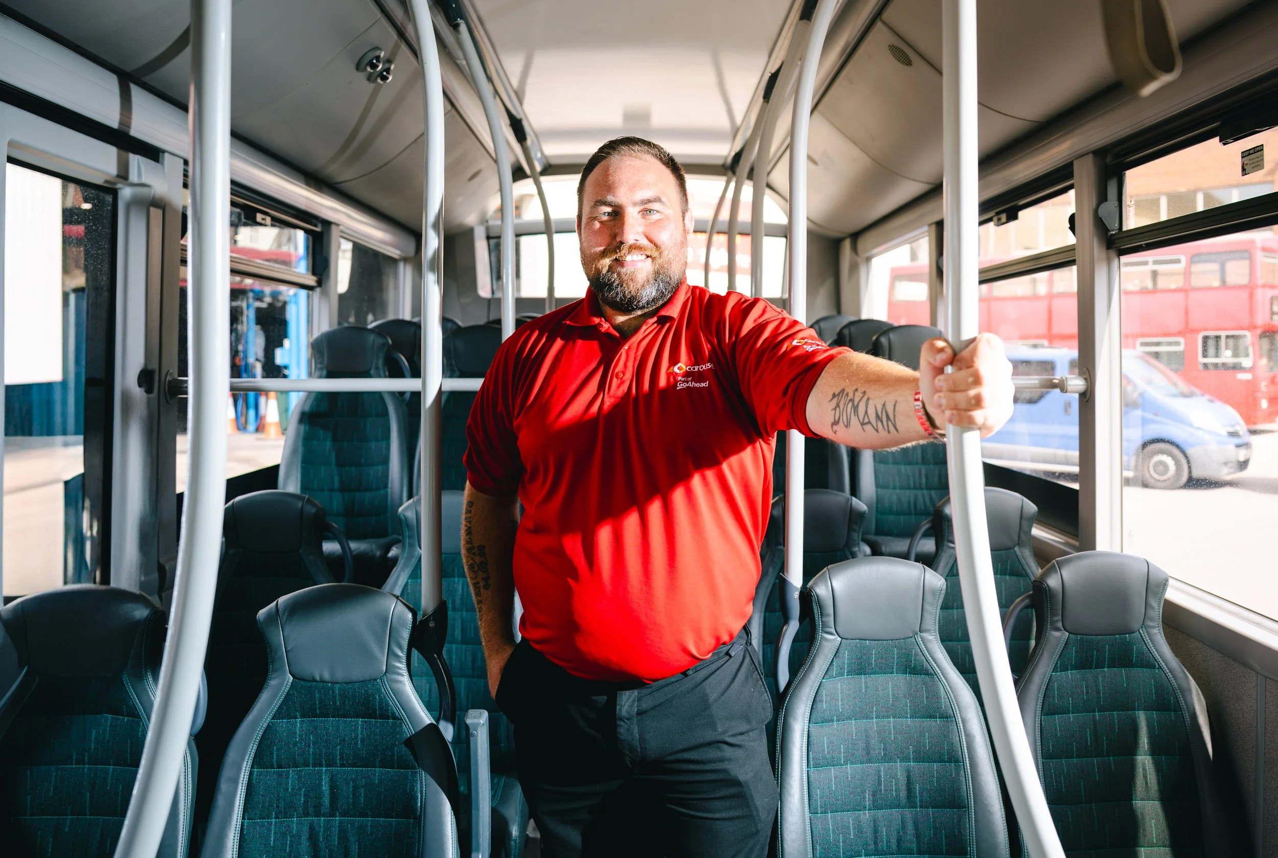 A man with a beard smiling and wearing a red uniform shirt inside a bus, standing next to a pole, with green and black bus seats visible behind him.
