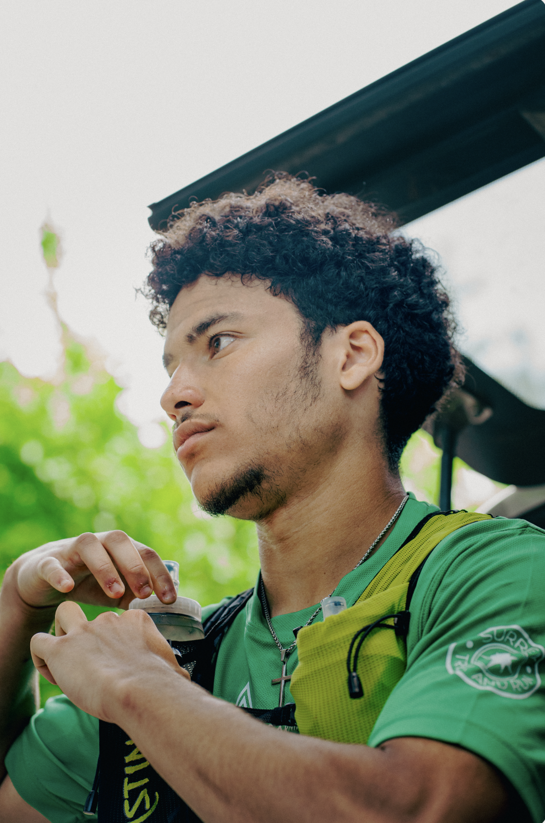 A young man with curly hair and a goatee in a green athletic shirt, holding a water bottle, outdoors with green trees in the background.