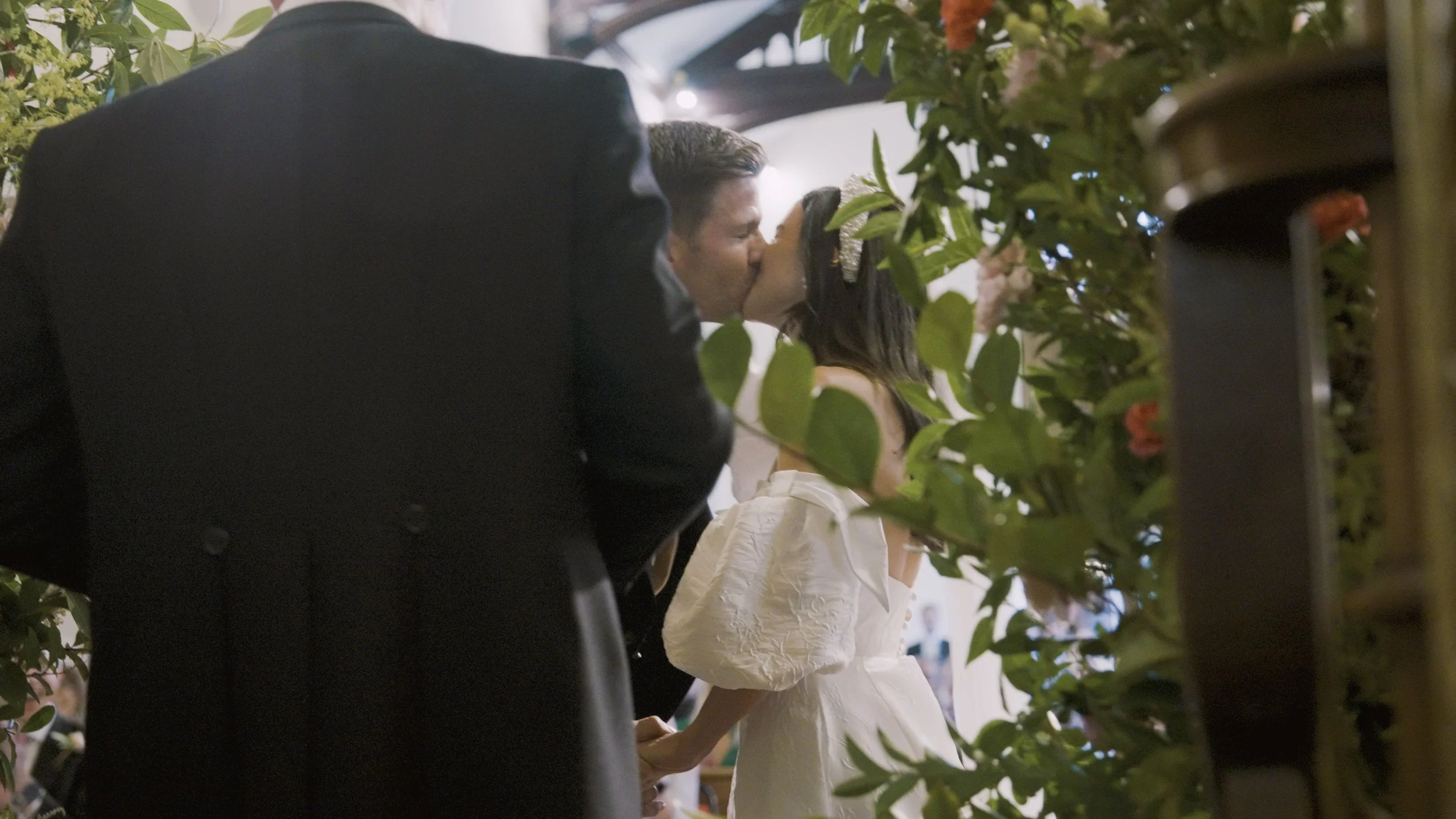 A couple shares a kiss during a wedding ceremony, with greenery surrounding them.