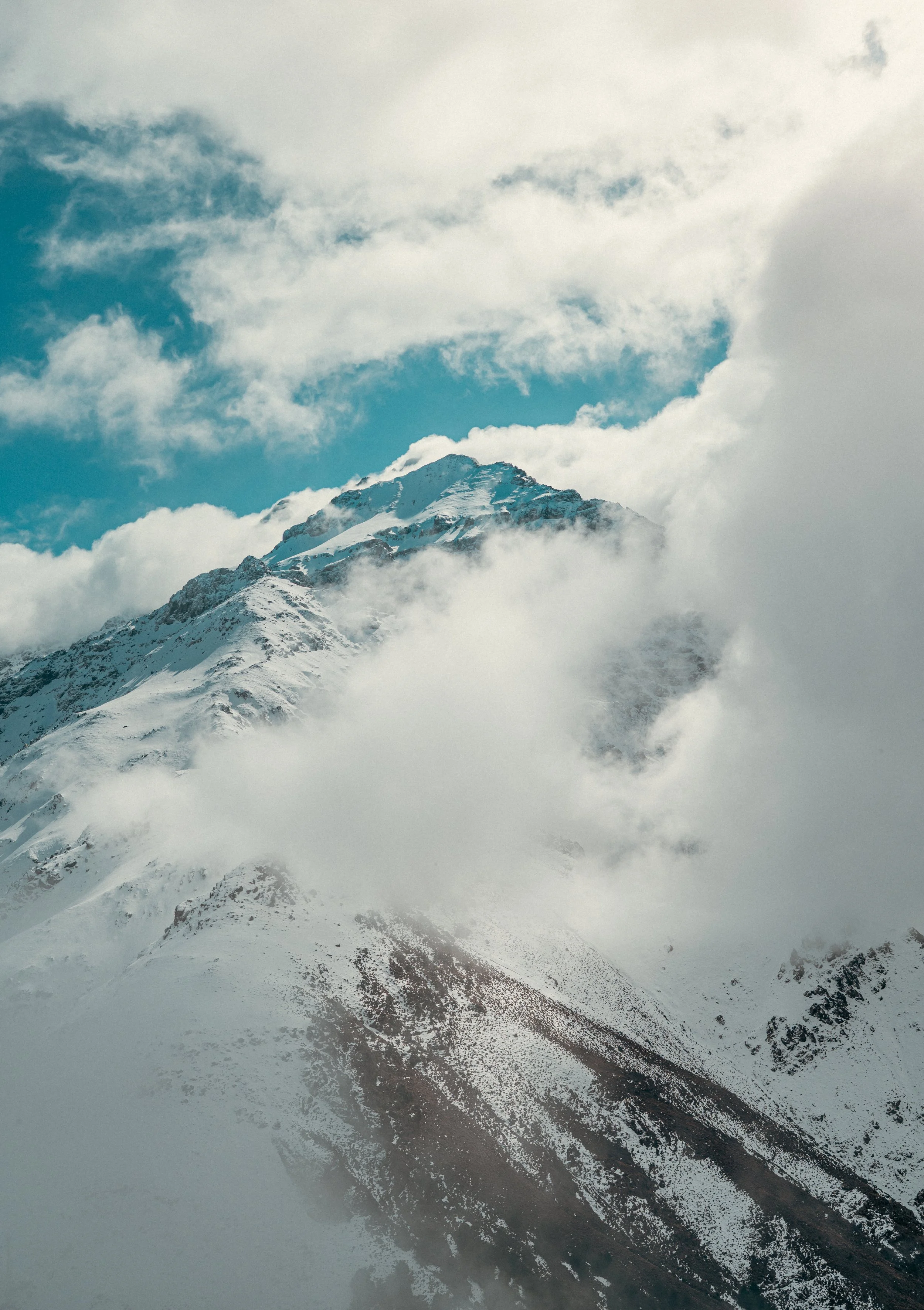 Snow-capped mountain peak partially obscured by clouds and fog, with rocky and snowy slopes and a mountain road at the base.