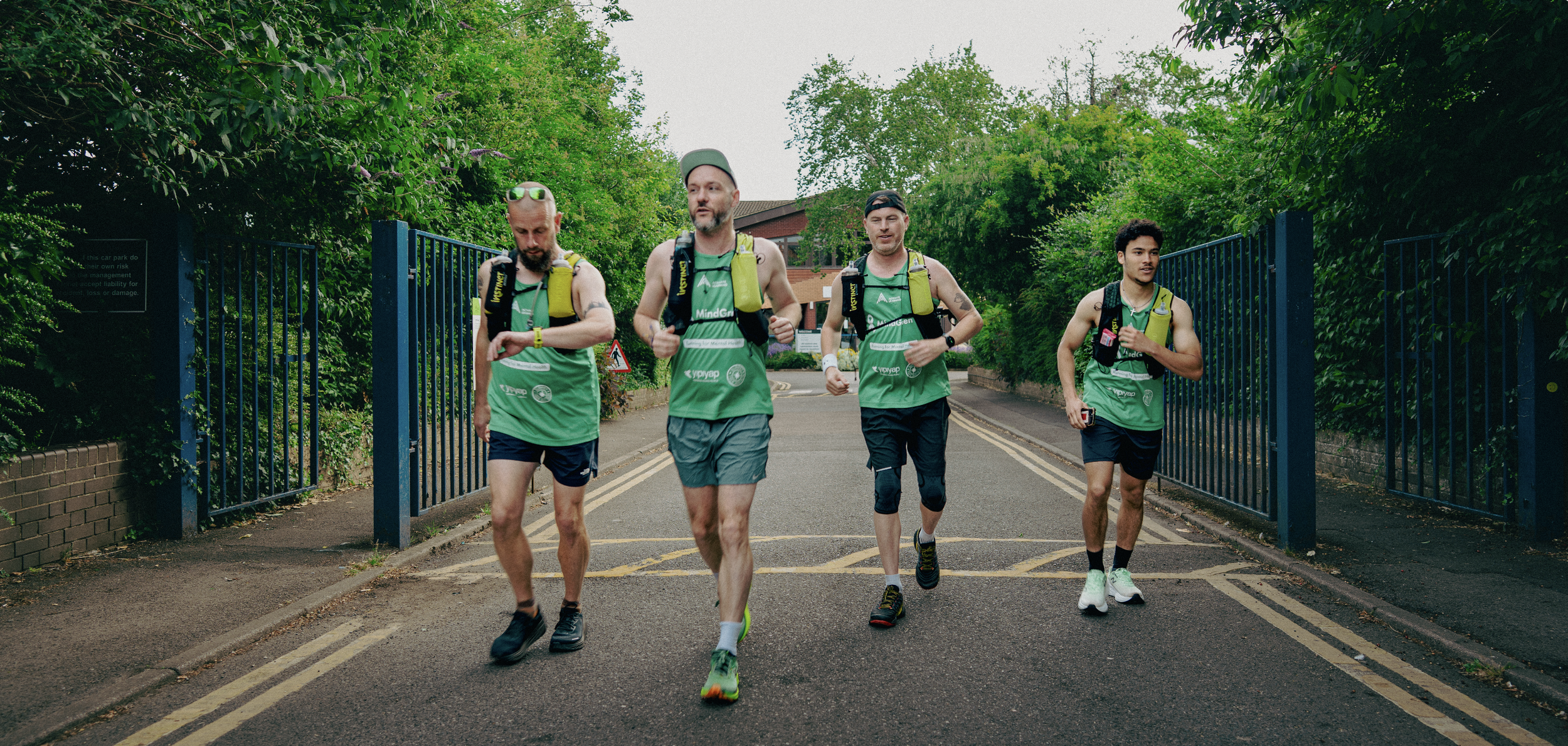 Four male runners in green shirts running on a street surrounded by green trees and bushes, with a blue gate and brick building in the background.