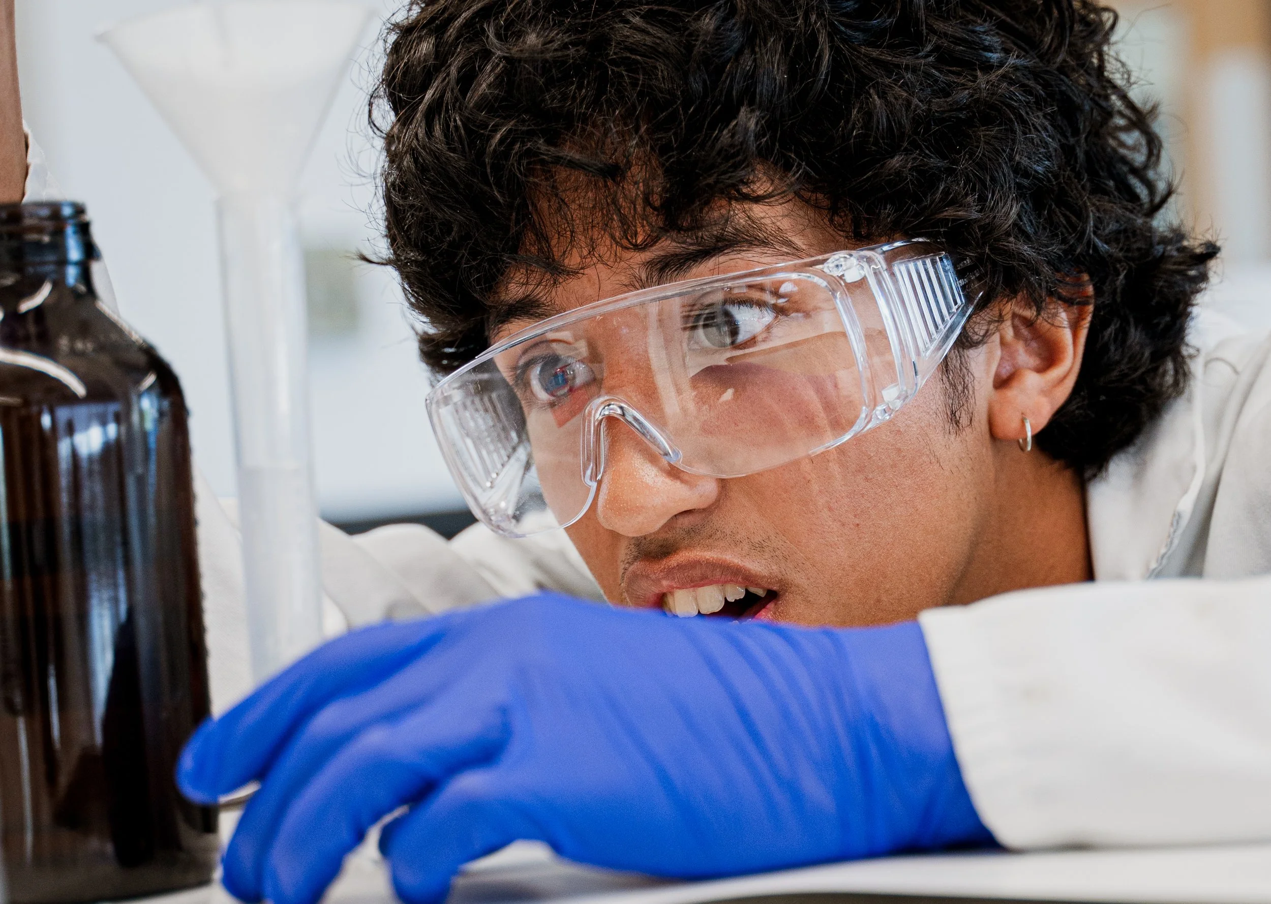 A scientist with curly dark hair, wearing safety goggles and blue gloves, is working in a laboratory, closely inspecting a sample or experiment.