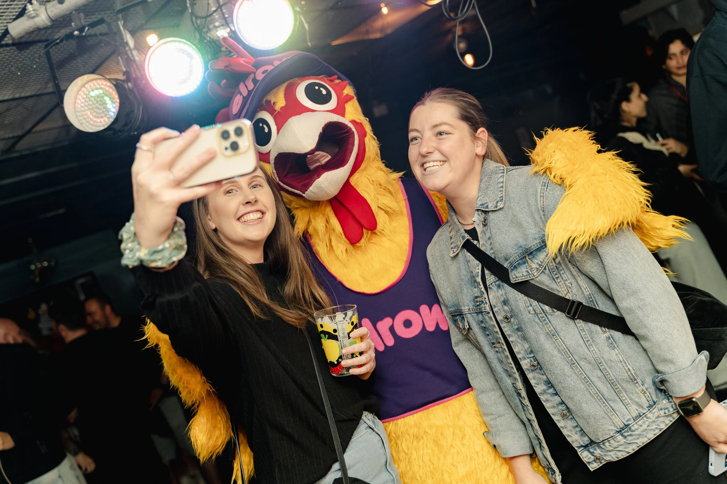 Two young women taking a selfie with a person in a colorful lion costume at a lively indoor event. The women are smiling, one holding a yellow drink, and the lion costume features bright yellow fur and a large, expressive face.