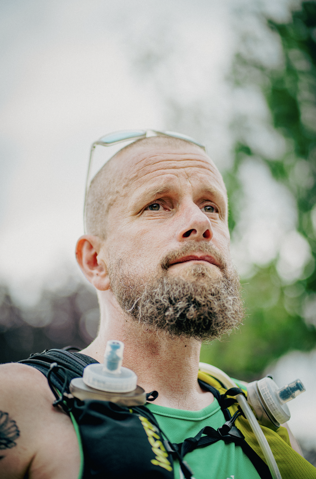 Close-up of a man with a bald head, glasses resting on his head, and a full beard, wearing a running vest with hydration gear, outdoors with blurred green foliage in the background.