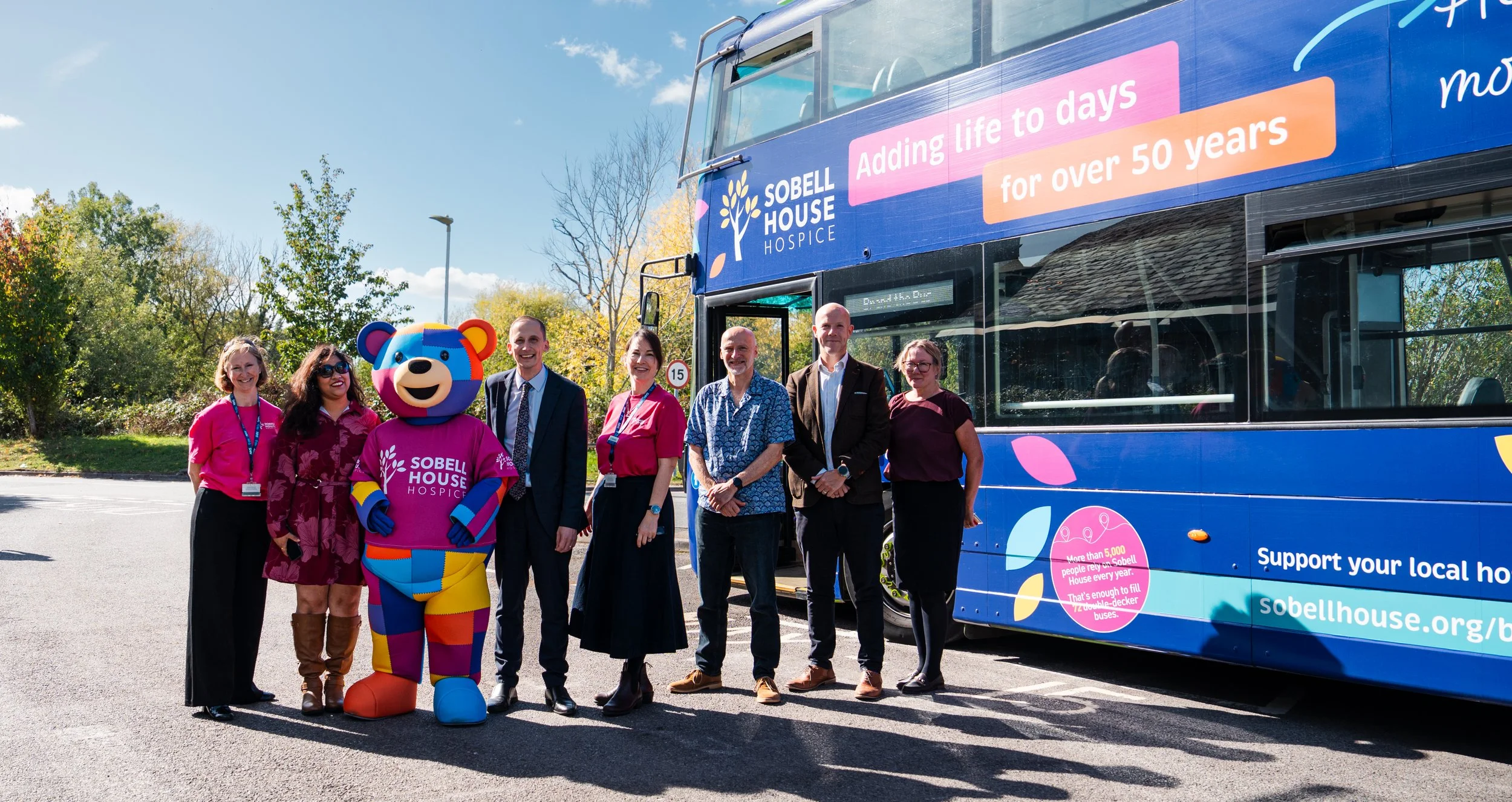 Group of people standing outdoors next to a blue bus with SOBELL HOUSE HOSPICE signage, including a mascot in a colorful bear costume, during daytime with trees and a clear sky in the background.