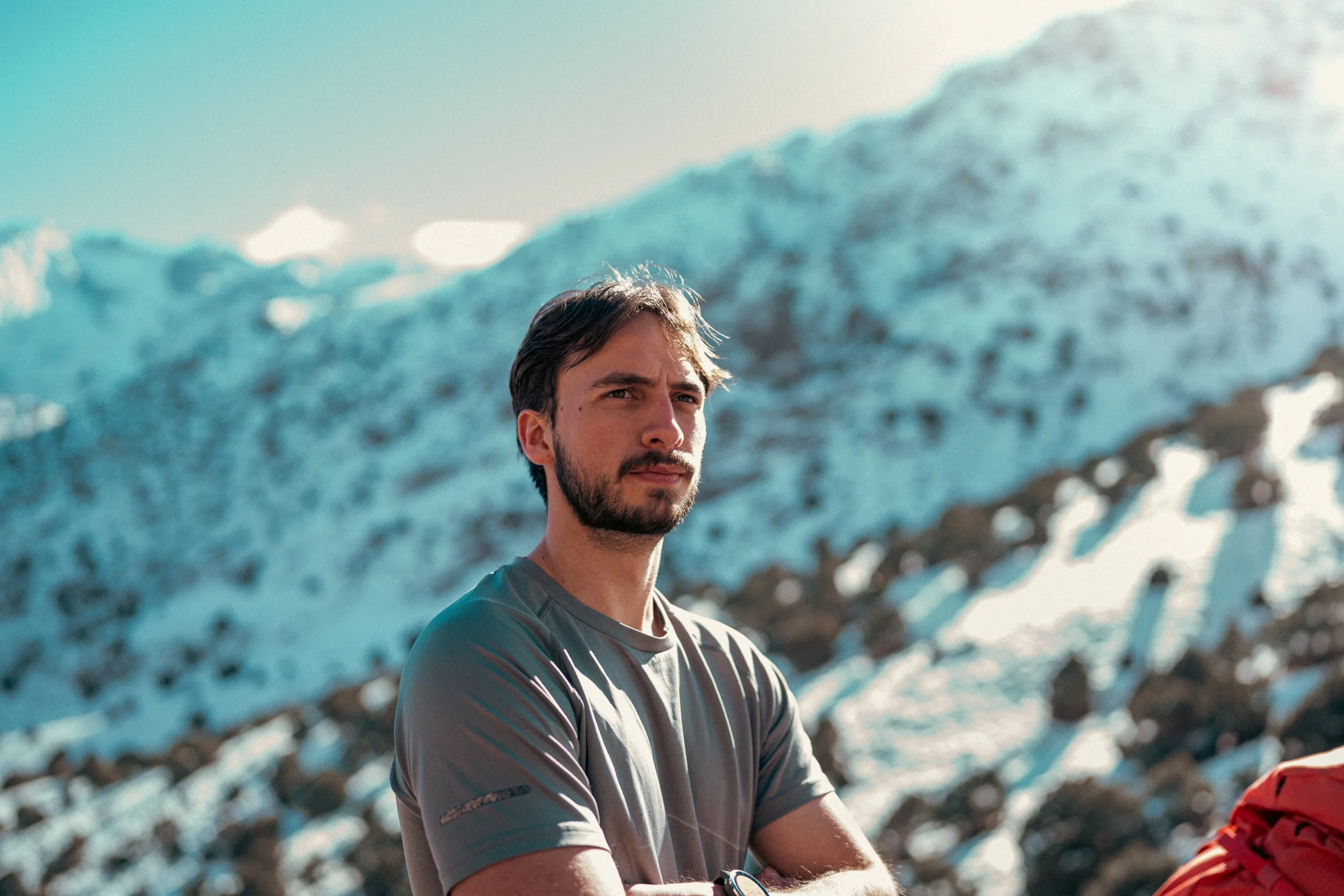 A man with dark hair and a beard in front of a large ice formation, wearing a gray shirt, with sunlight illuminating the scene.
