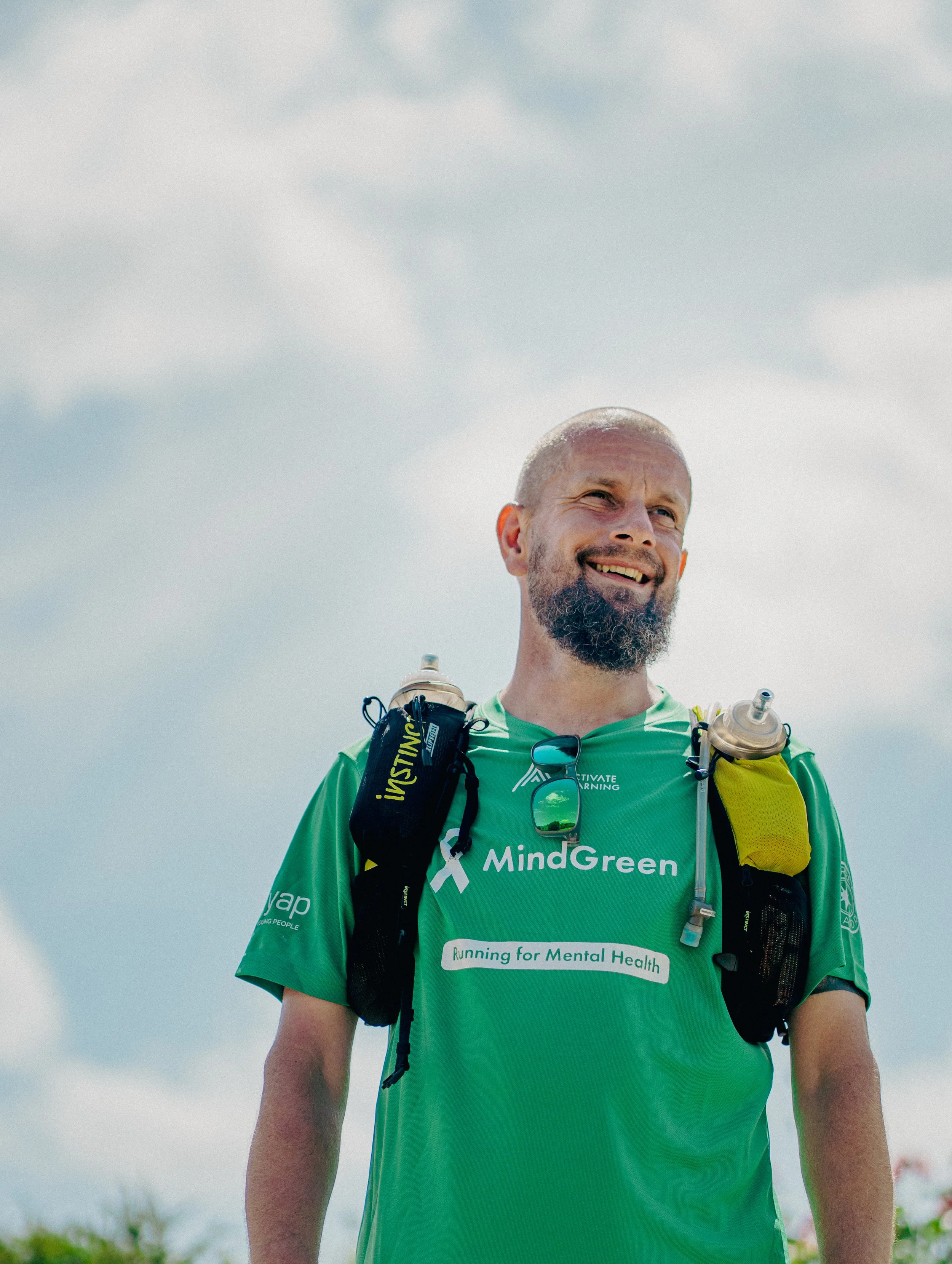 A smiling man wearing a green running shirt with the text 'Running for Mental Health' and a white ribbon logo, carrying a hydration pack, outdoors on a cloudy day.