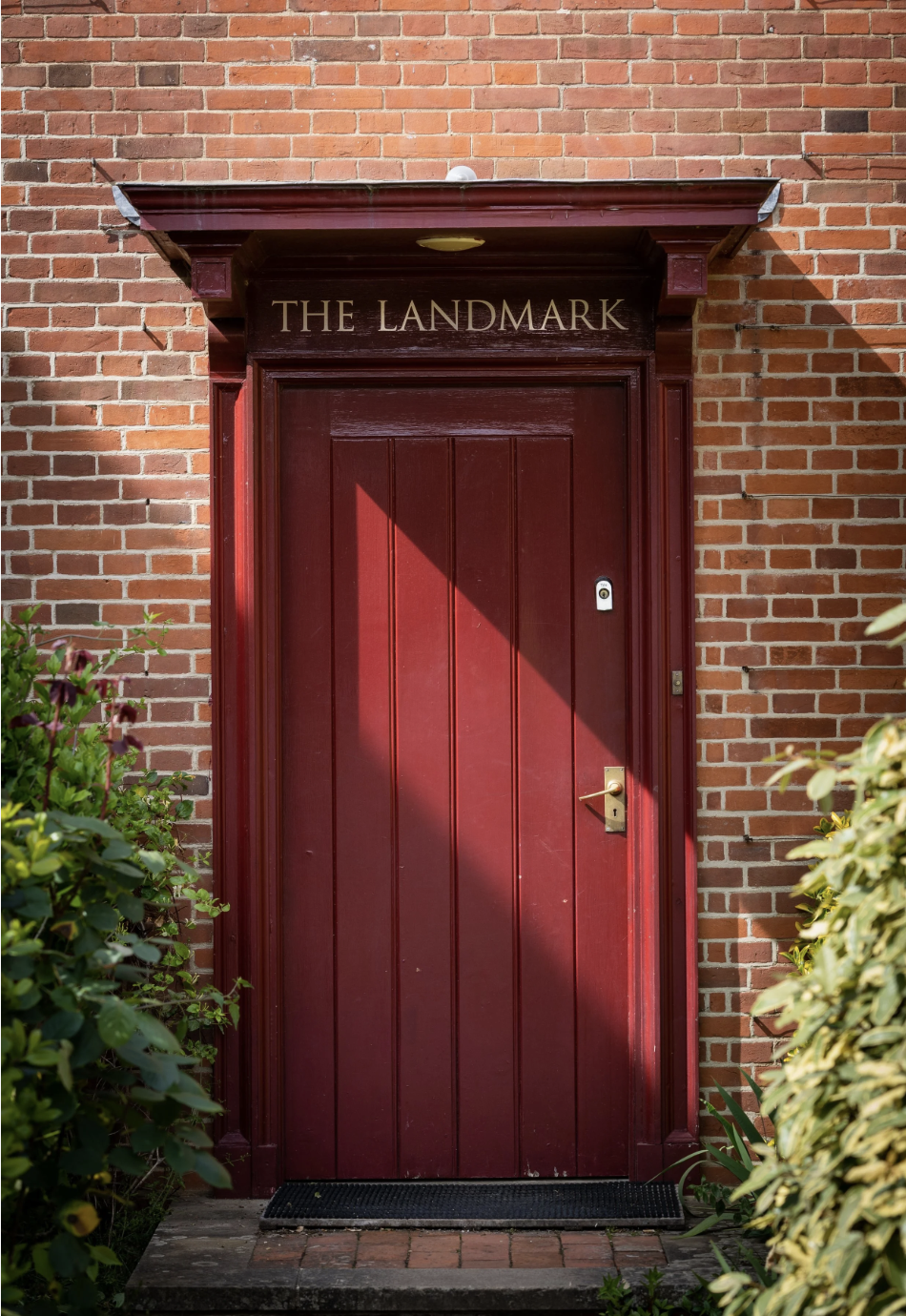Red front door with a black doormat, surrounded by green bushes, on a brick building, with a sign above that reads 'The Landmark'.