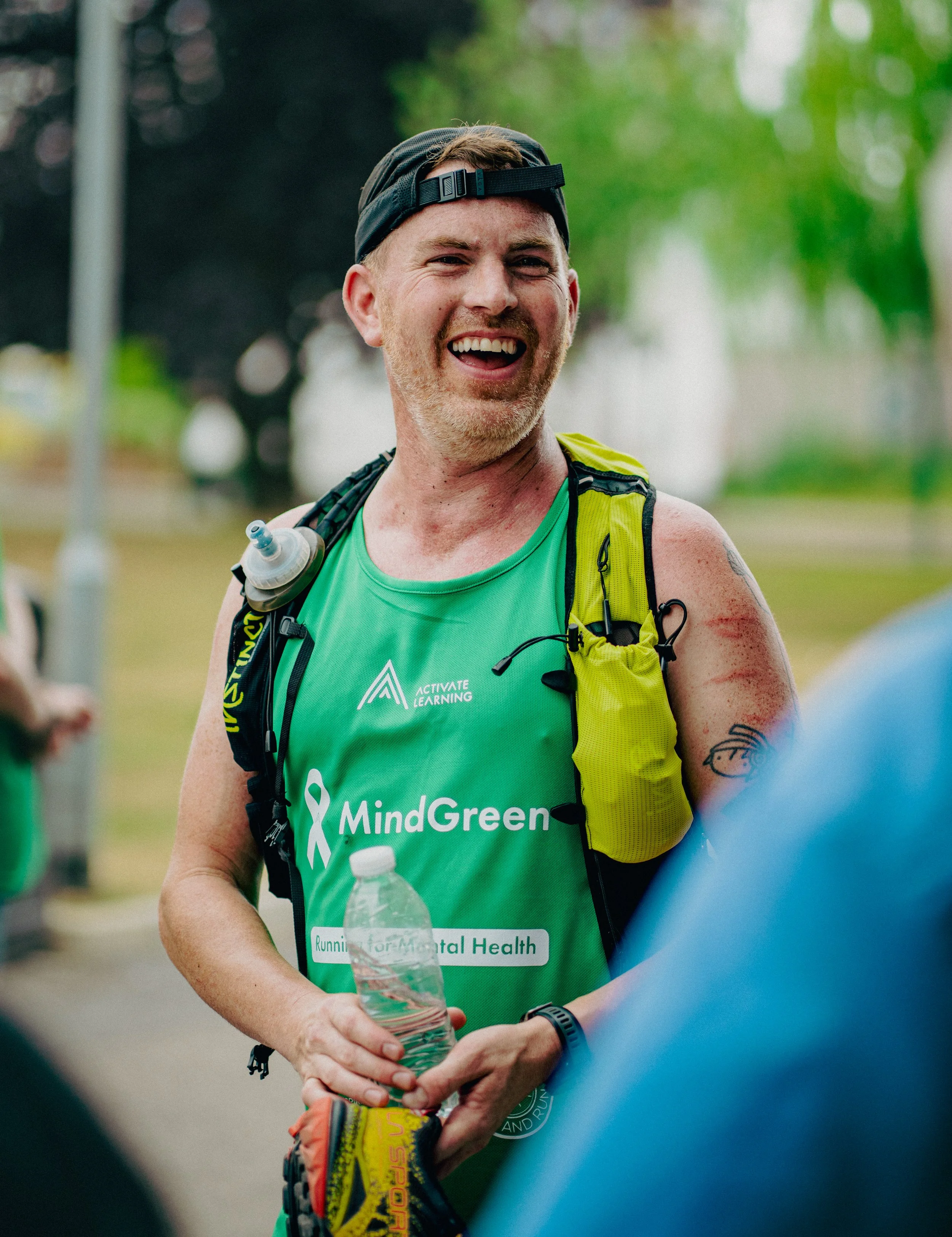 A man smiling and wearing a black cap backwards, a green tank top with a logo and text, and a yellow backpack, holding a water bottle, outdoors with blurred trees and people in the background.