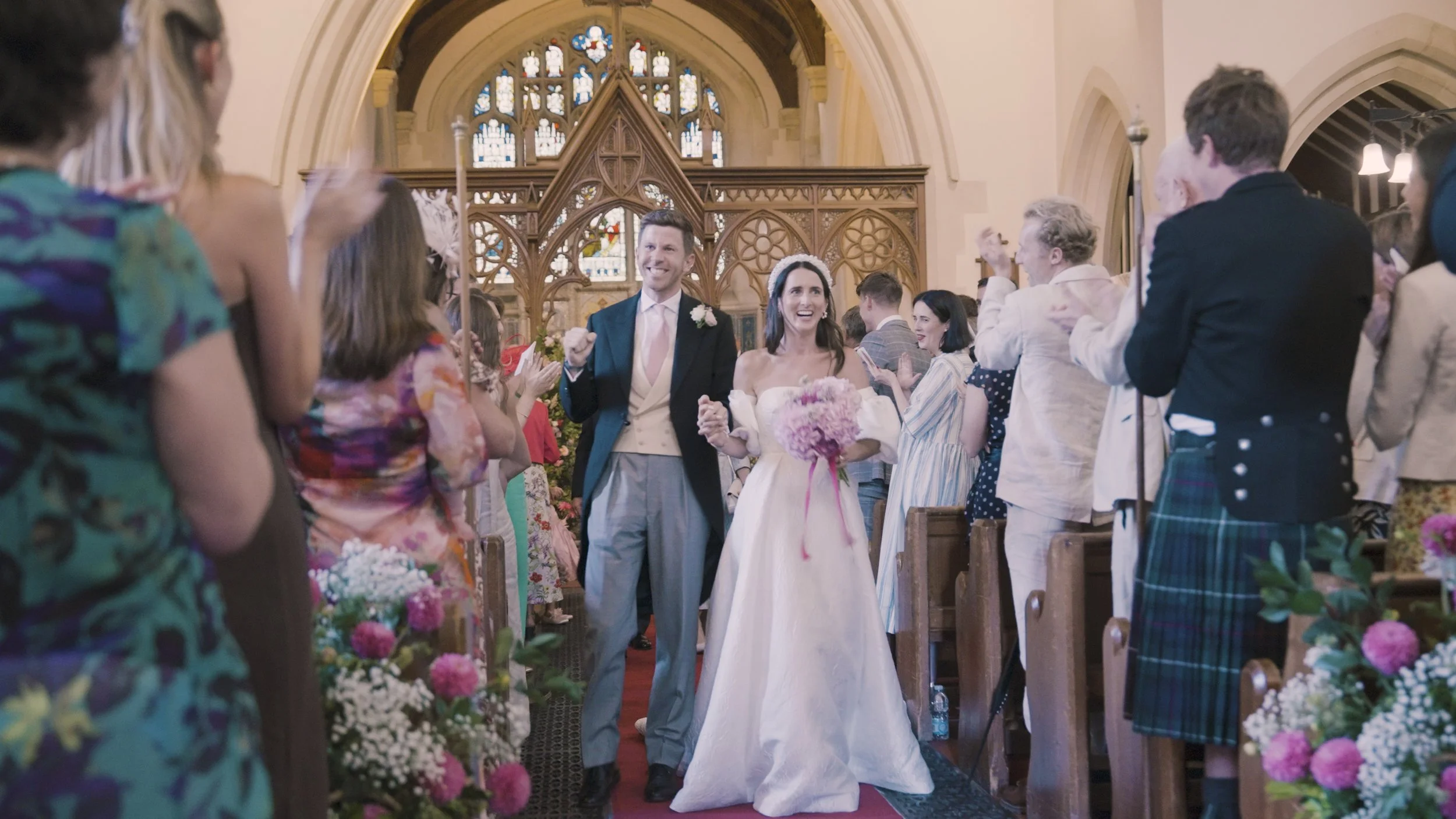 A newly married couple walking down the aisle in a church decorated with flowers, surrounded by smiling guests clapping and celebrating.
