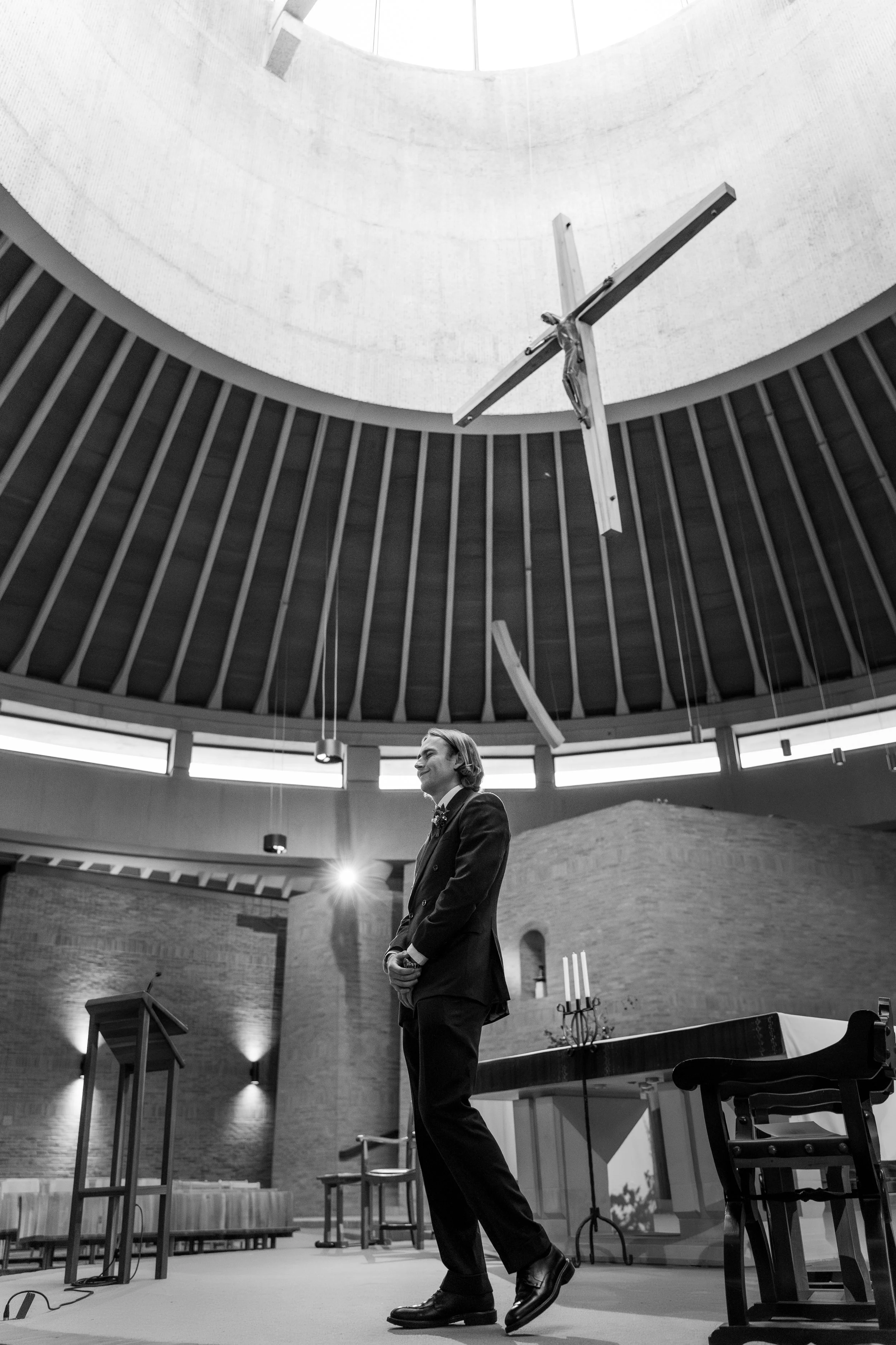 A man dressed in formal attire standing inside a church, with a large crucifix hanging from the ceiling above him.