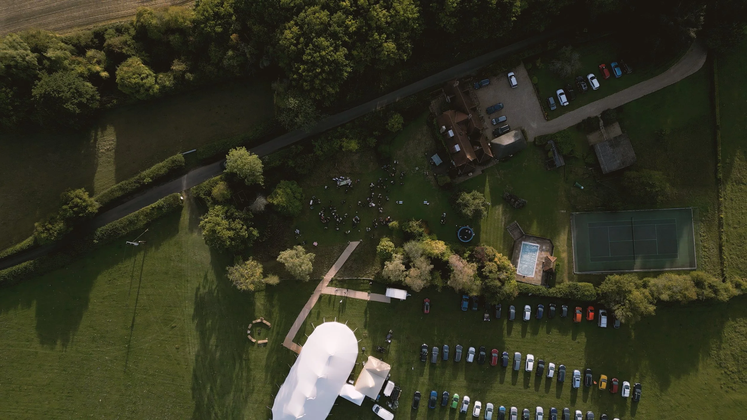 An aerial view of a large outdoor gathering with many cars parked around a grassy area, a group of people under a tent, and a tennis court, surrounded by trees and fields.