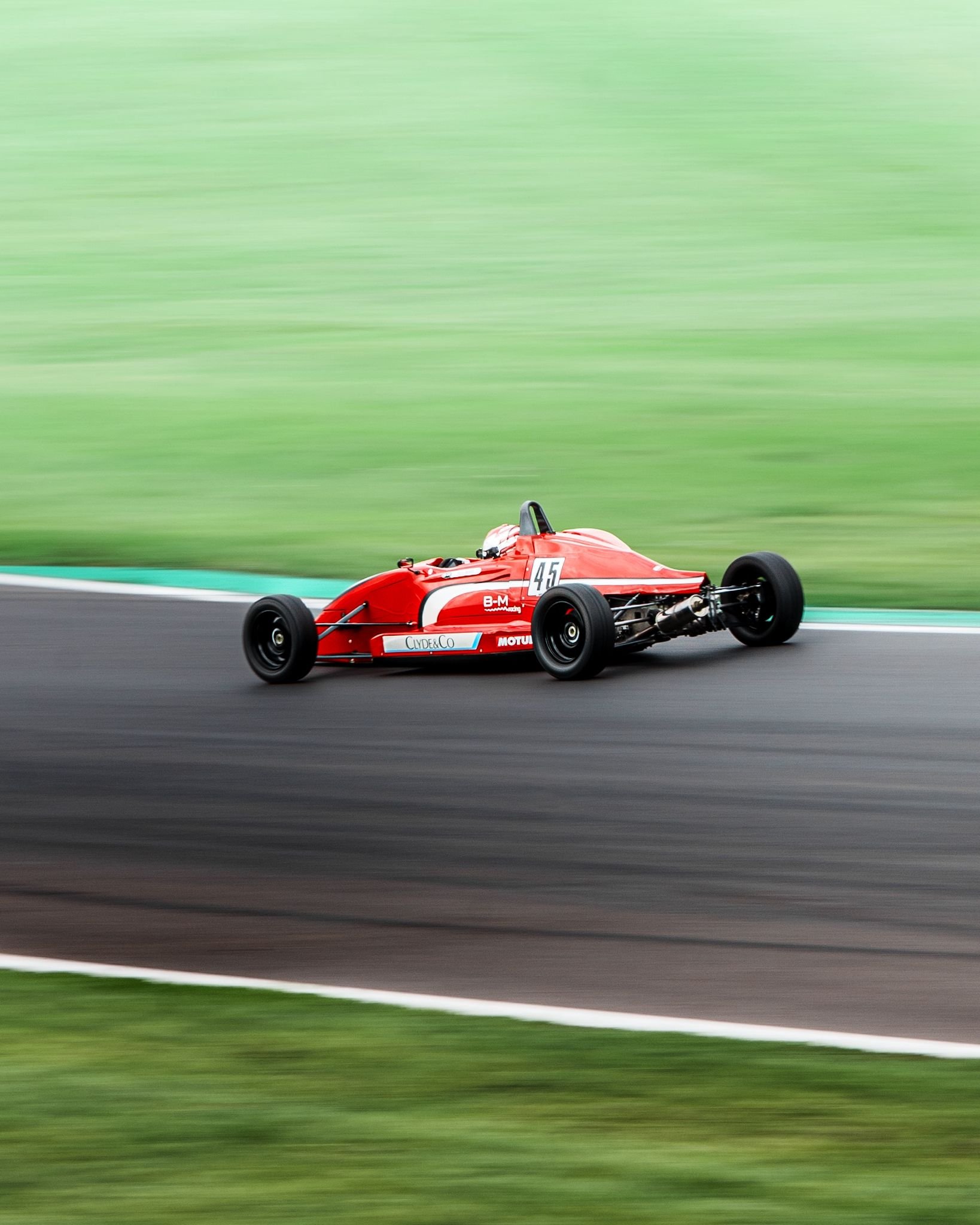 Red vintage race car with the number 45 racing on a track, surrounded by a blurred green grassy background.