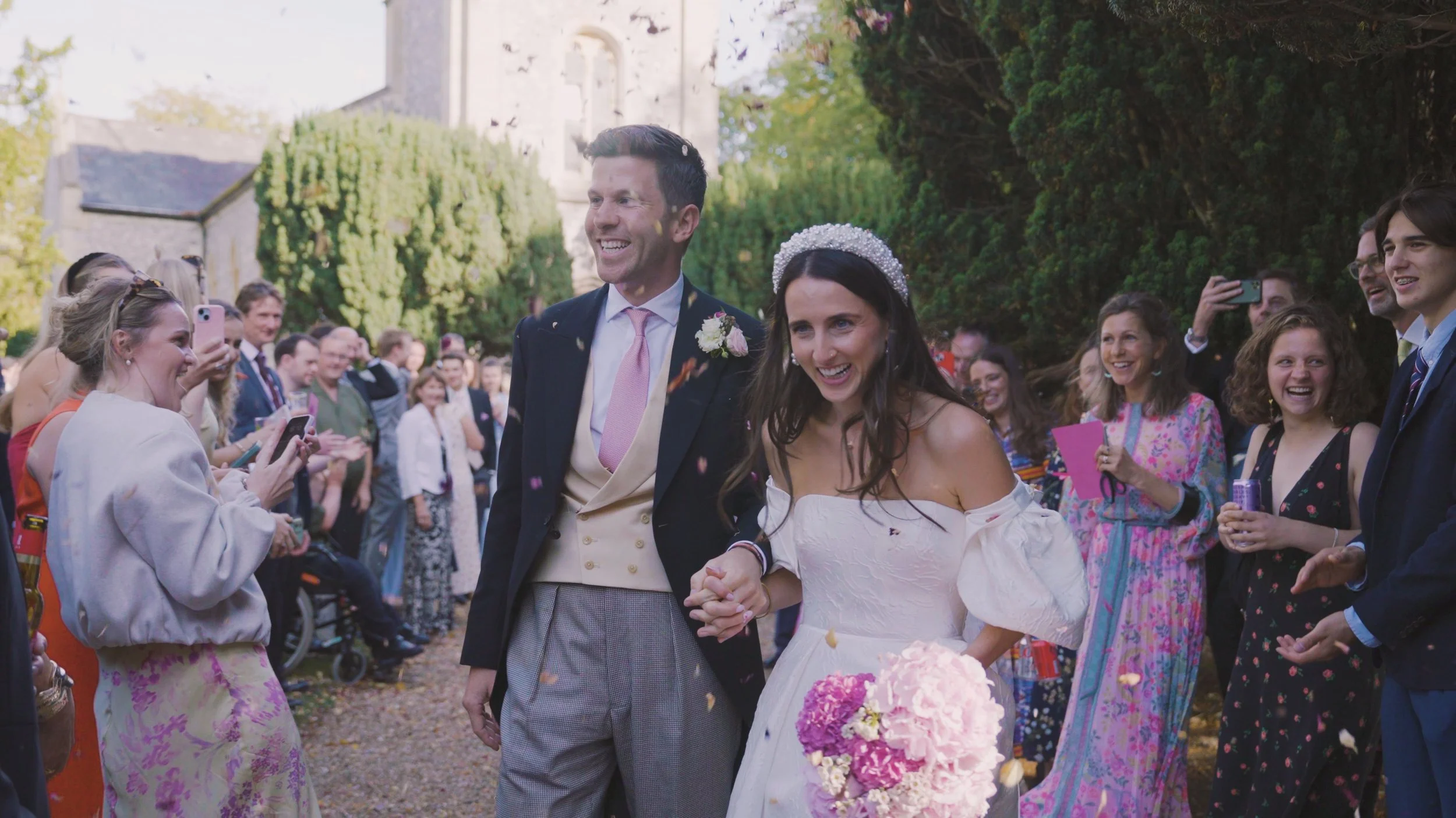 A newly married couple holding hands and smiling while walking outdoors during their wedding celebration. The groom wears a dark suit with a pink tie and boutonniere. The bride wears a white wedding dress and a pearl headpiece. Guests surround them, 