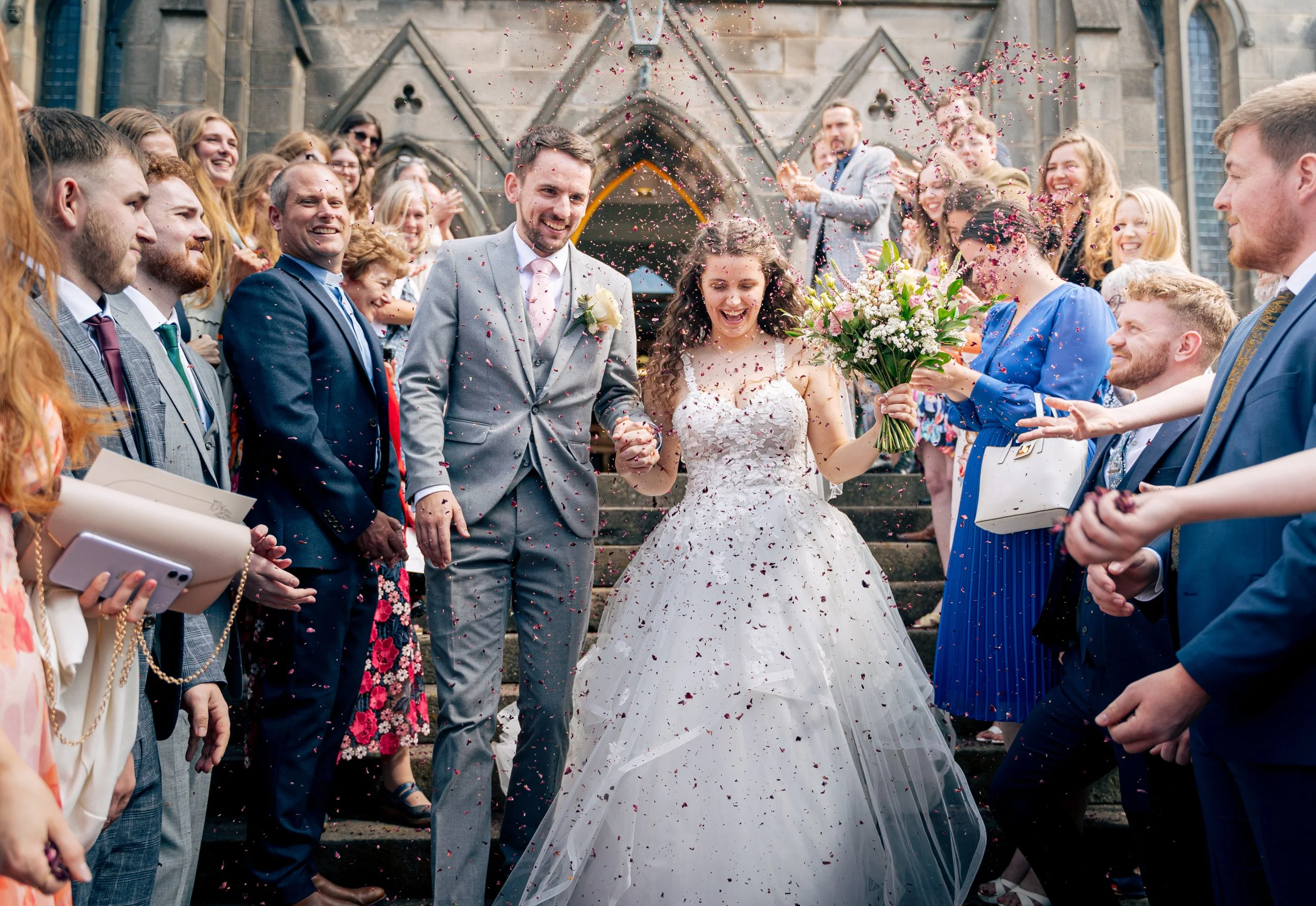 A wedding celebration outside a church with the bride and groom surrounded by guests throwing flower petals. The bride is holding a bouquet and the groom is in a gray suit with a pink tie and boutonniere.
