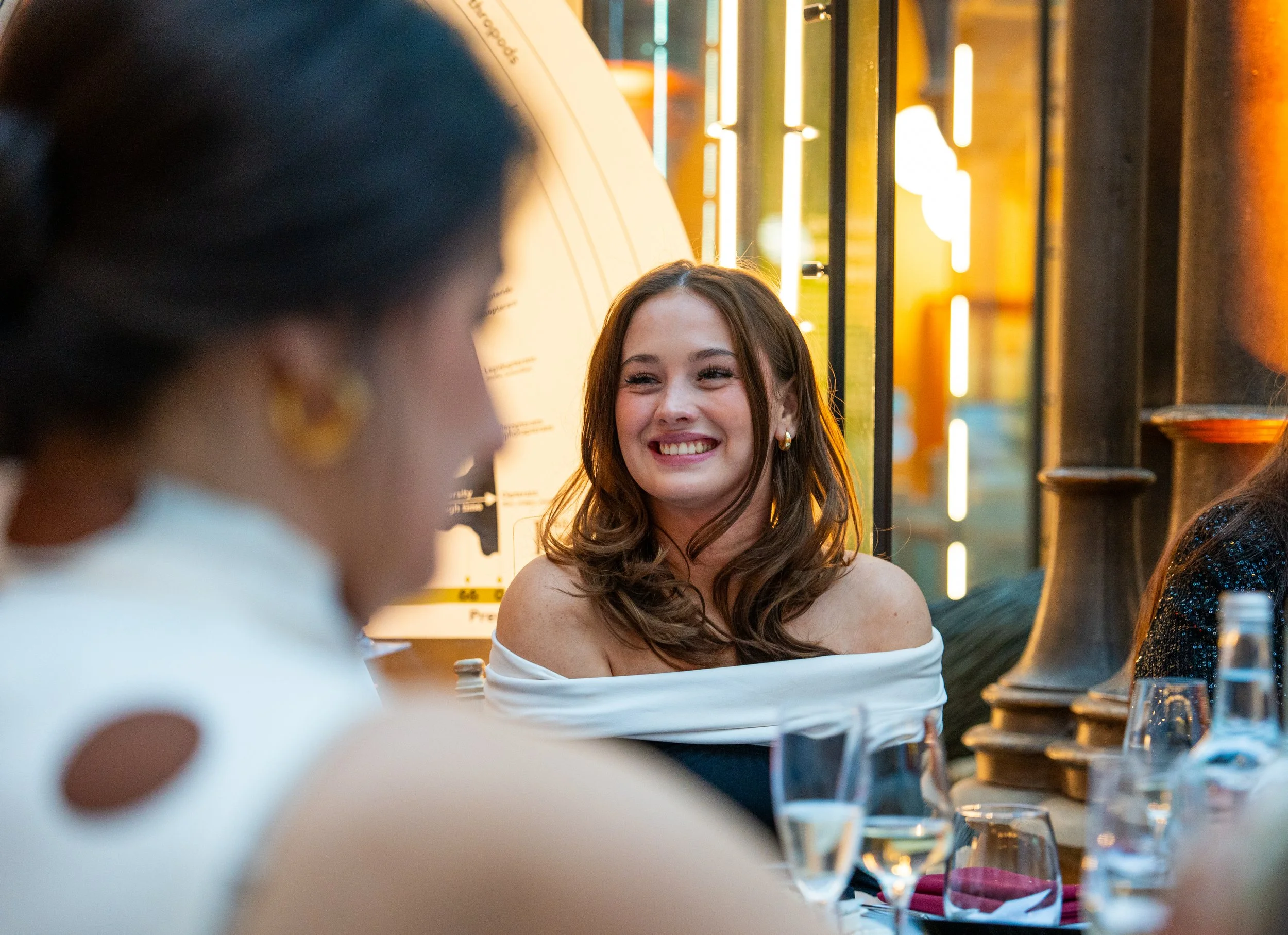 Smiling woman in off-the-shoulder white dress at a social gathering or dinner party with wine glasses and bottles on table.