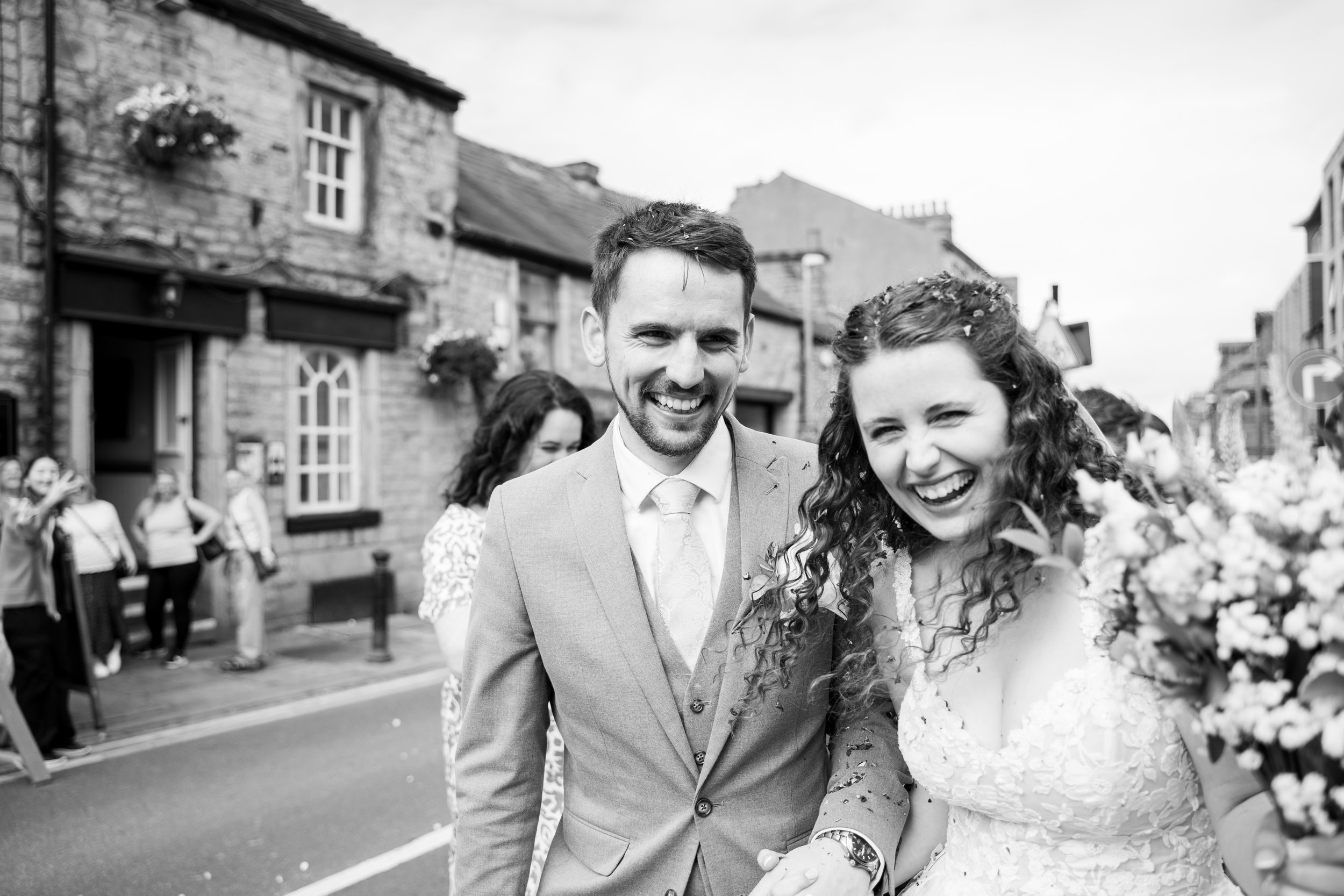 A joyful couple, possibly newlyweds, walking outdoors on a city street, smiling and holding hands. The man is in a suit, and the woman is in a lace dress with flowers in her hair.