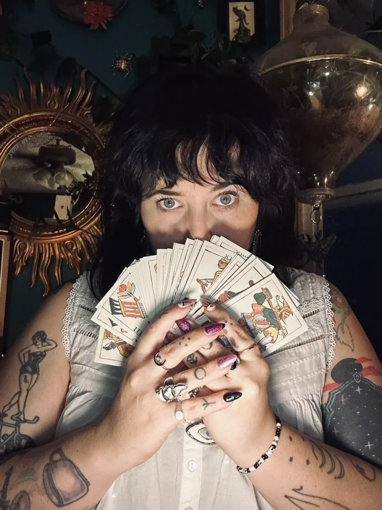 Woman with tattoos holding a fan of tarot cards, indoors with a large decorative mirror and a glass lamp in the background.