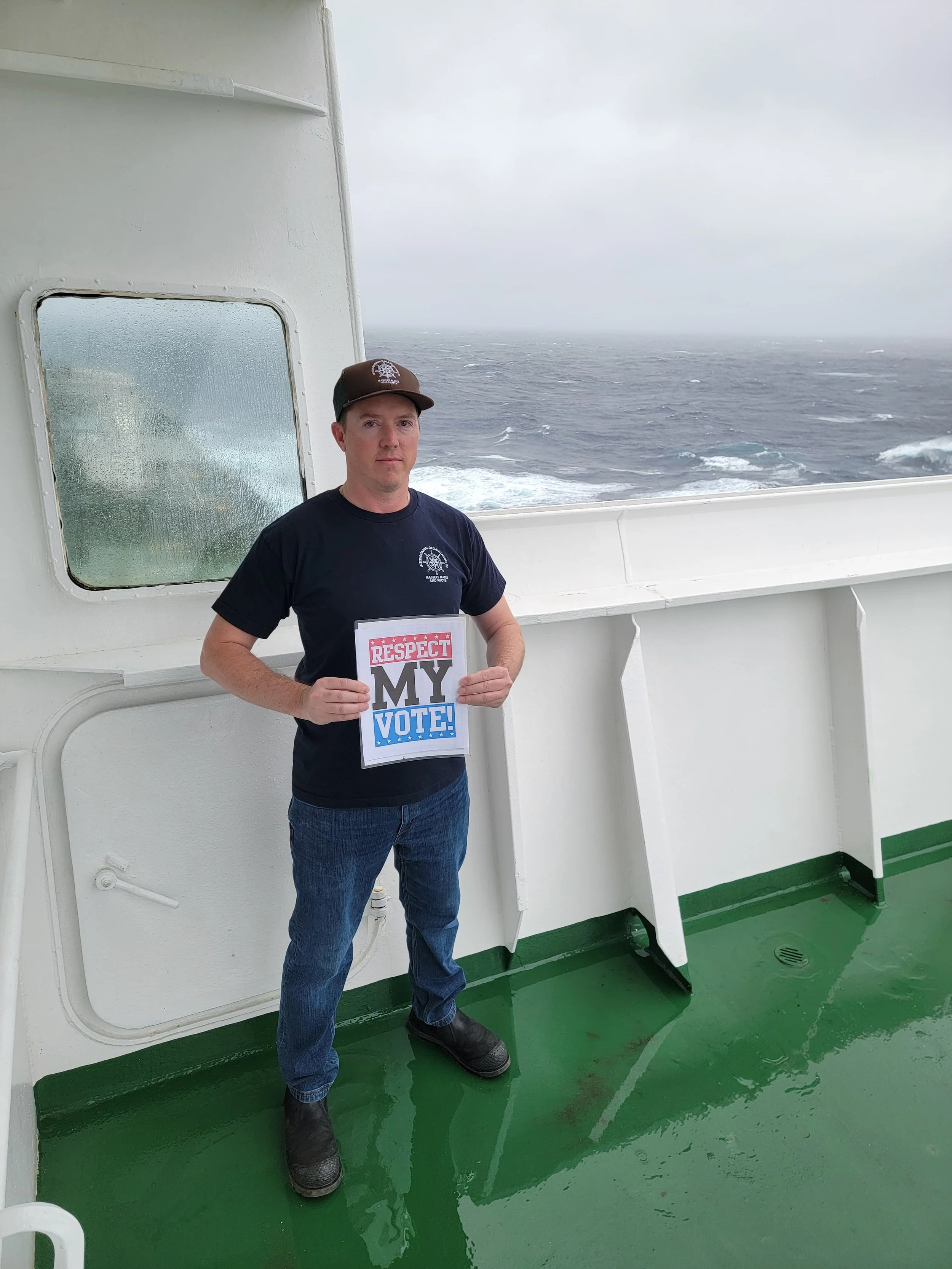 ATC Mate aboard a ship holding a “Respect My Vote!” sign with rough ocean waves behind him