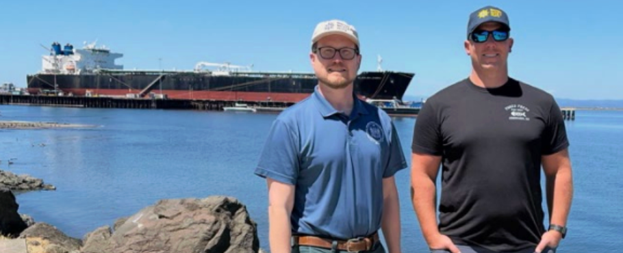 MM&P Secretary Treasurer Lars Turner and ATC Mate posing by a rocky waterfront with a large tanker ship in the background.