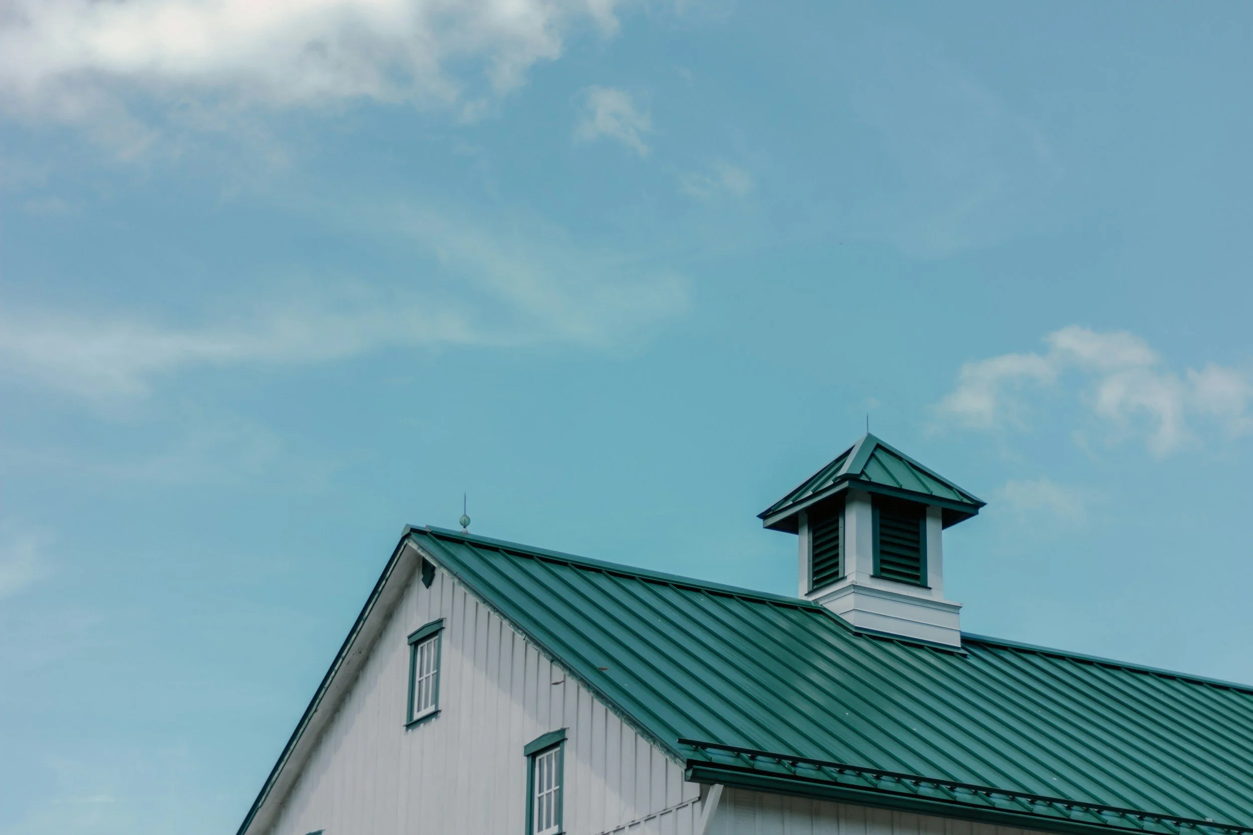 A white house with a green metal roof, a small tower with louvered vents, and a partly cloudy sky in the background.