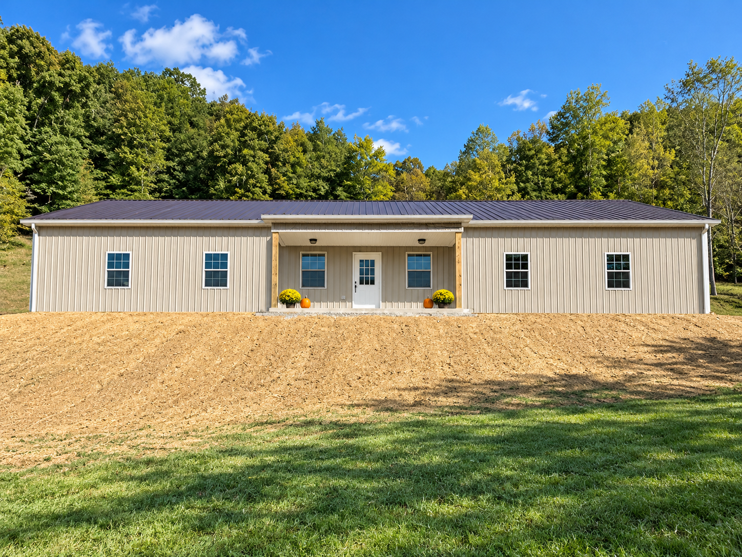 A newly built beige house with a dark metal roof, four windows on the front, and a white door centered under a small porch with wooden columns. There are two yellow potted plants and two pumpkins on the porch. The house is on a sloped dirt yard with 