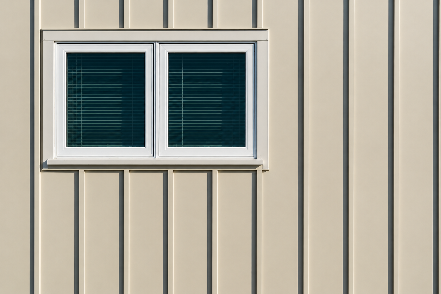 Exterior wall of a building with beige siding and a white-framed window with closed blinds.