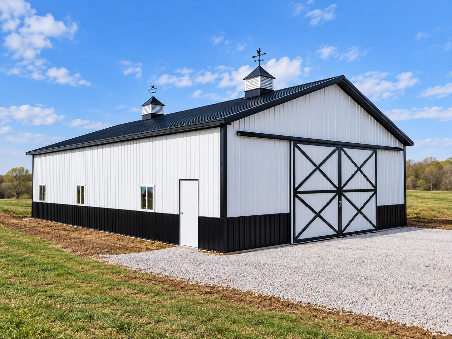 A white and black barn with a metal roof and two cupolas on top, set in a rural field under a blue sky with clouds.