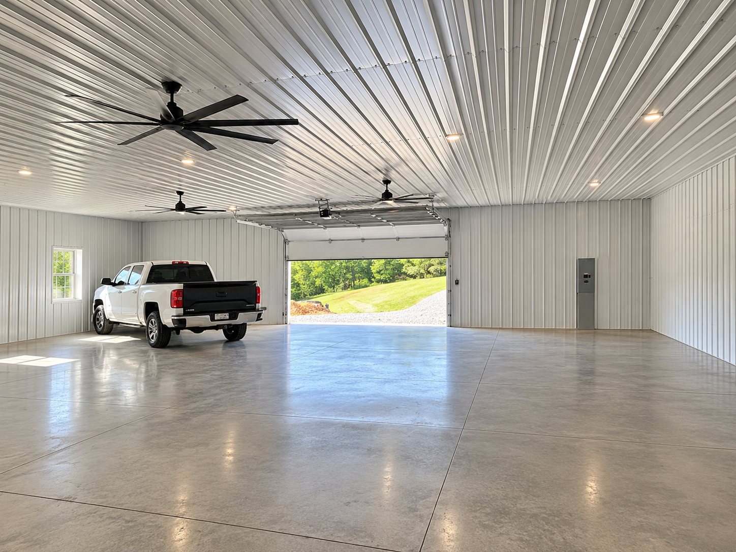 A spacious, clean, empty garage with a white pickup truck parked inside. The garage has a metal ceiling with ceiling fans and recessed lighting, a large open garage door revealing green grass and trees outside, a small window on the left wall, and an