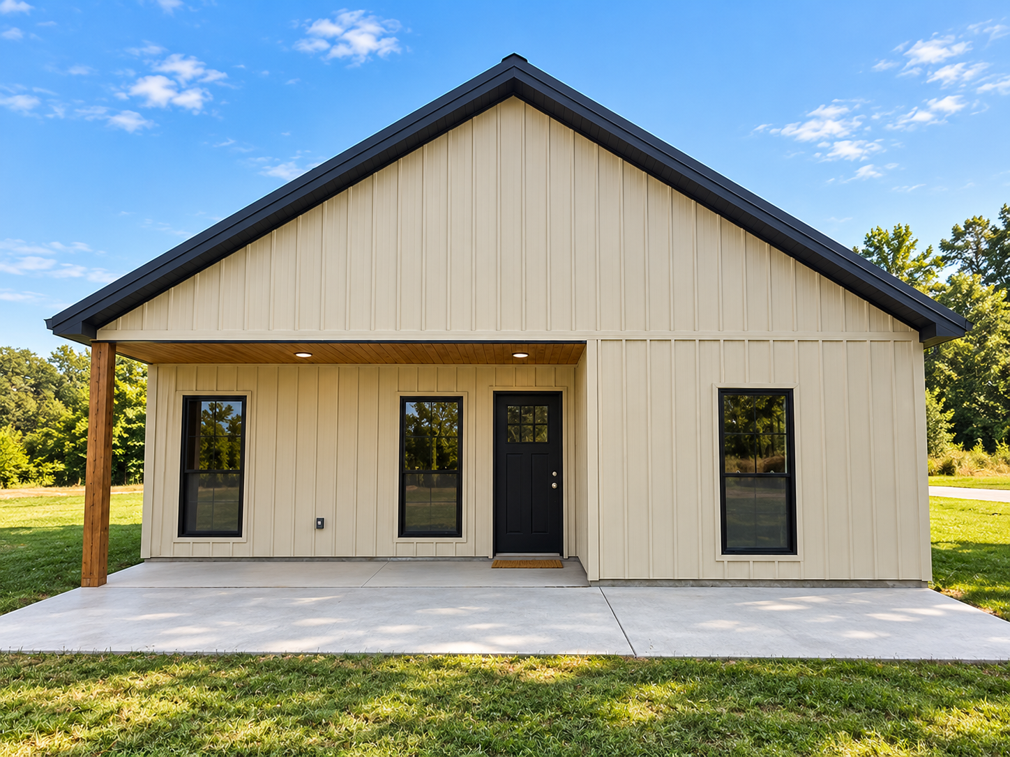 A modern, beige-colored house with black windows and a black front door, with a small porch and a concrete patio, surrounded by grass and trees under a blue sky with some clouds.
