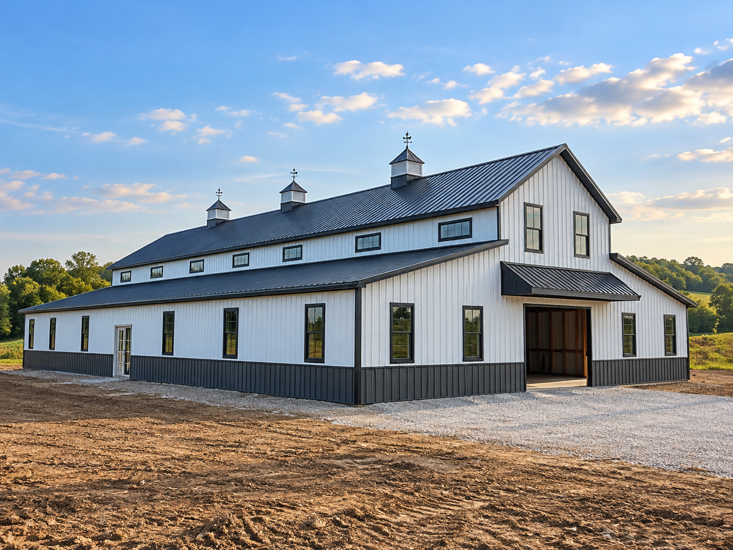 Large white barn with black trim and a metal roof, situated in a rural area under a blue sky with scattered clouds.