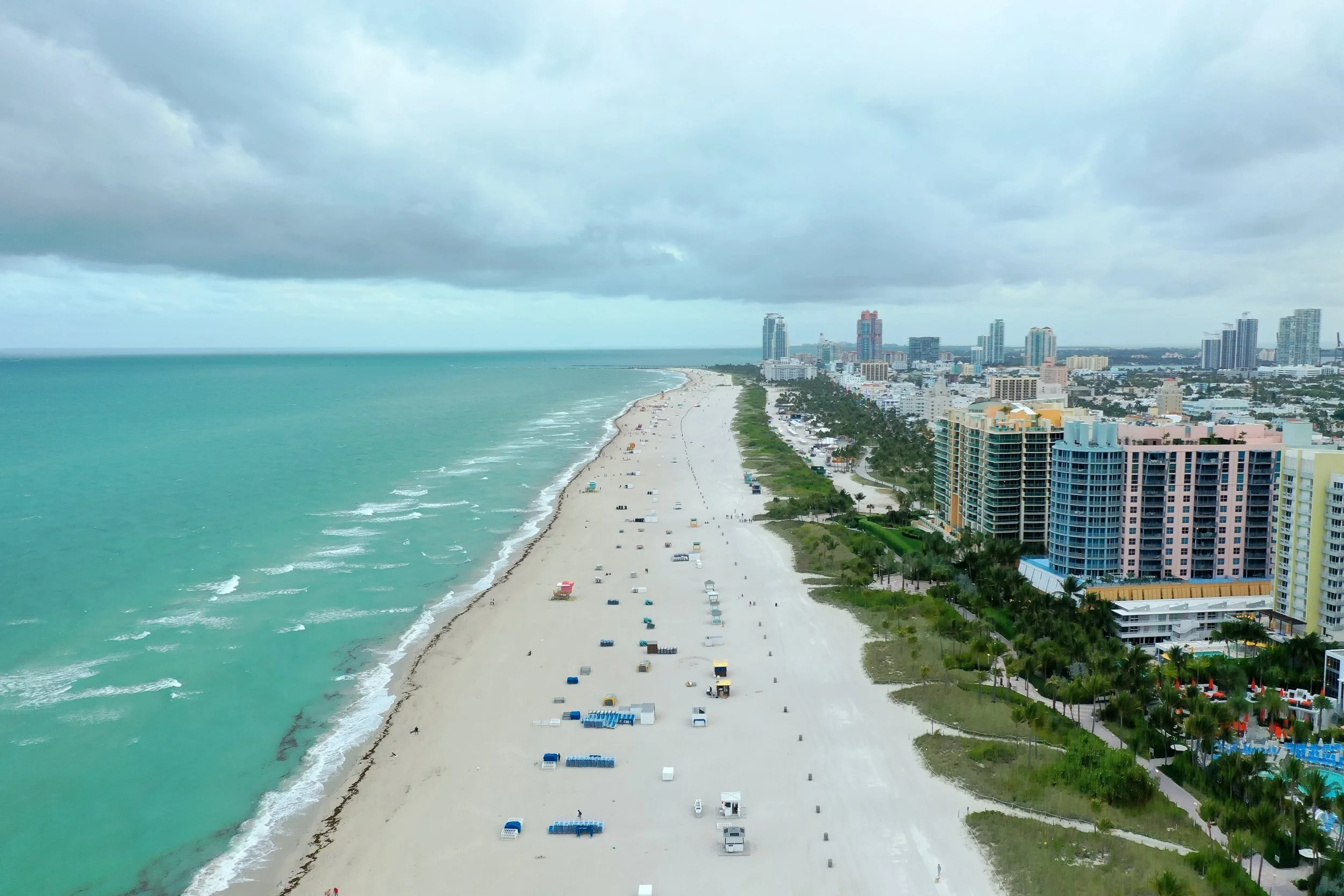 A city skyline with high-rise buildings along a beach under a cloudy sky, showing a shoreline with beach chairs and umbrellas and a row of colorful high-rises and hotels.