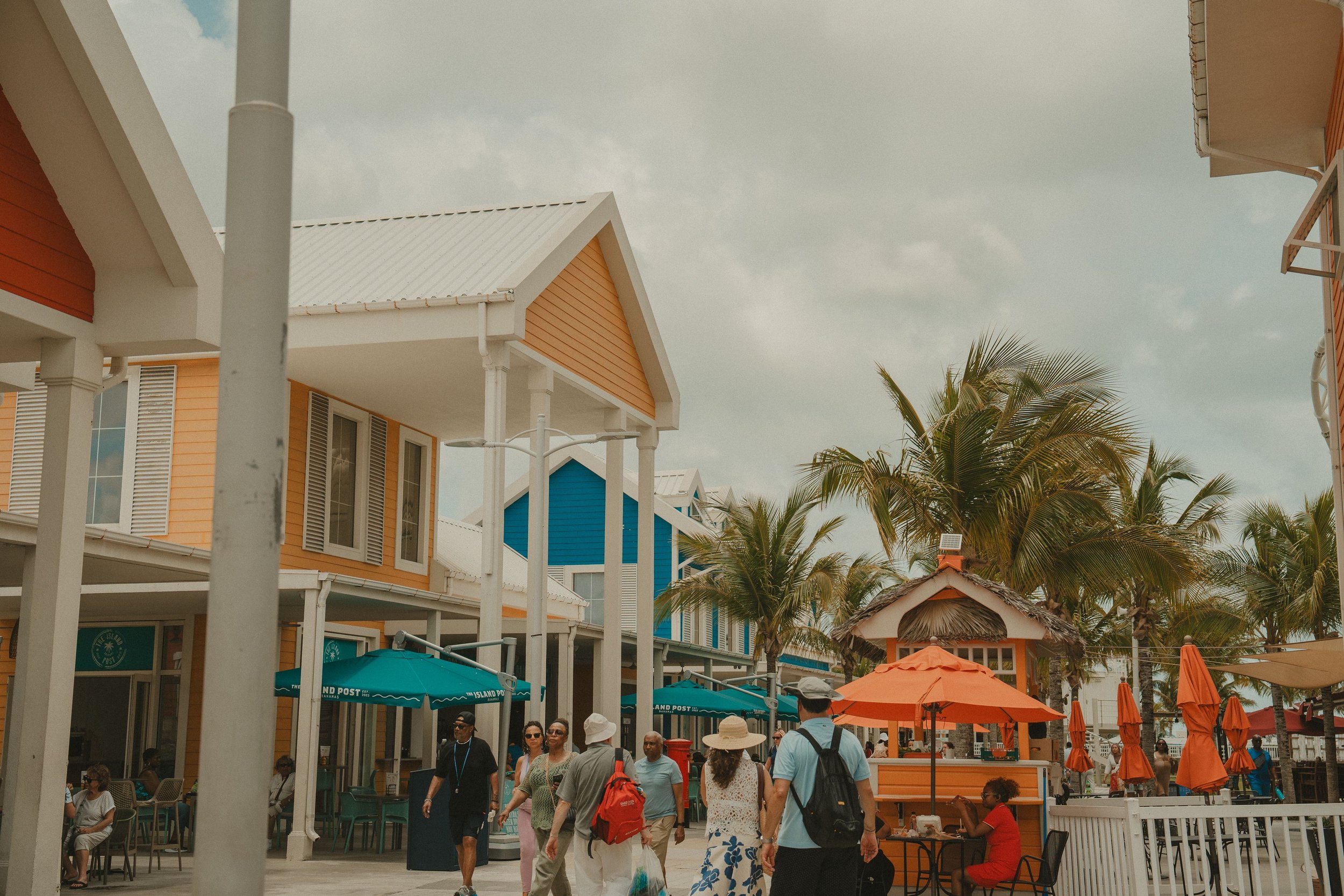 People walking and sitting at shops and cafes with colorful houses, palm trees, umbrellas, and a cloudy sky in a busy outdoor shopping area.