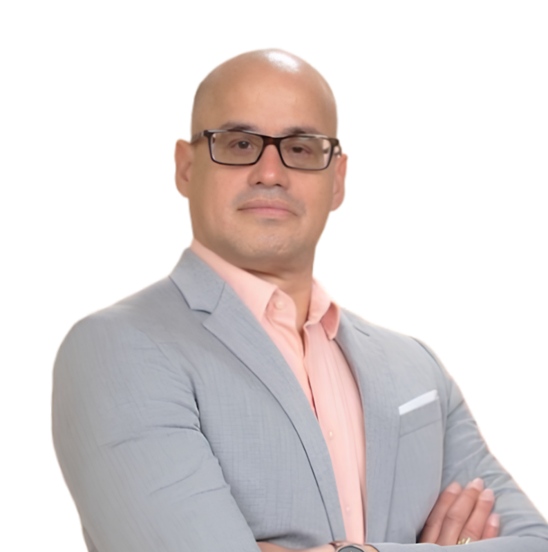 A man in a light gray suit with a pink shirt, wearing glasses, standing with arms crossed against a plain white background.