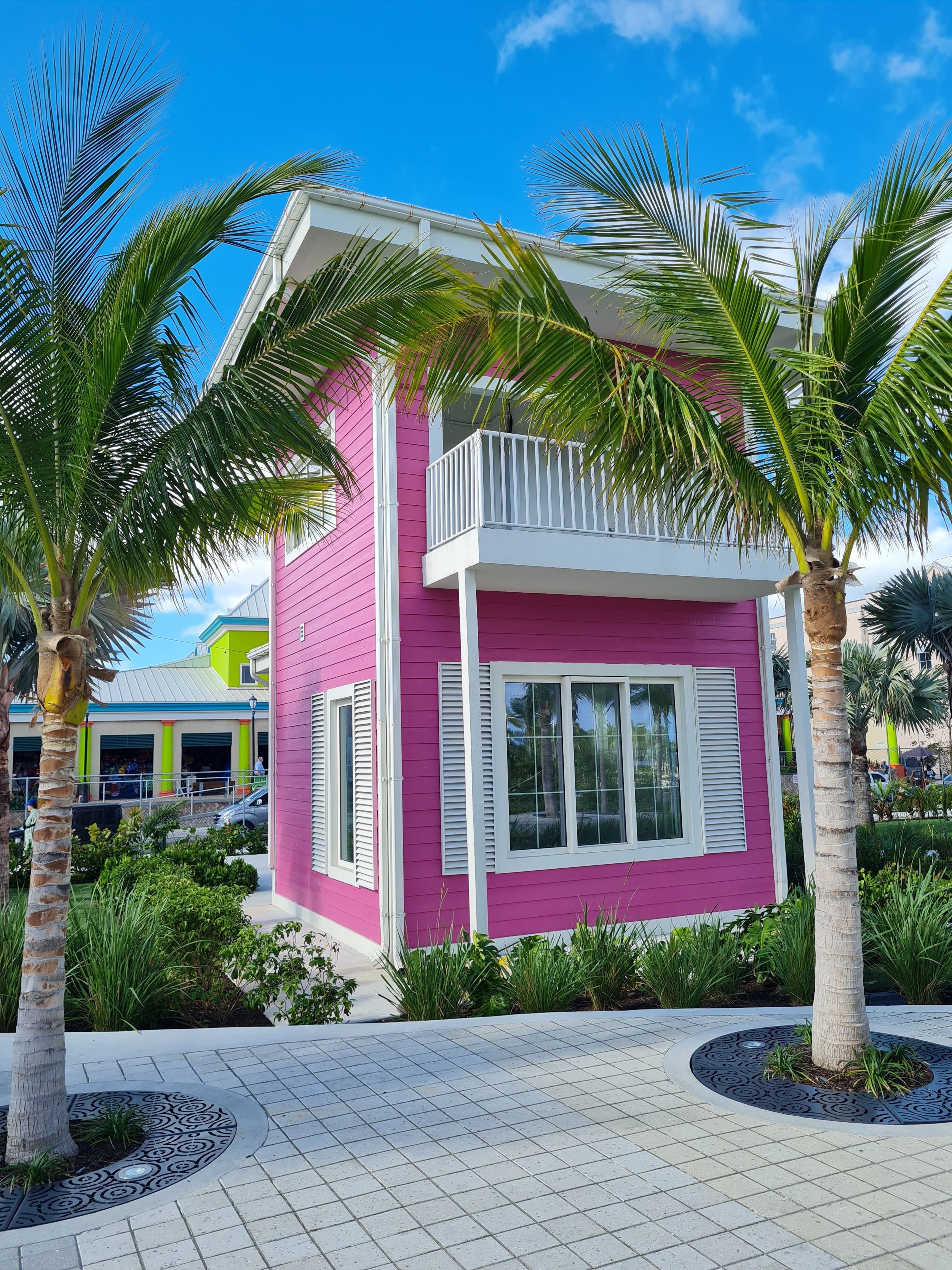 A pink house with white trim and a balcony, surrounded by palm trees and greenery, on a bright sunny day with a blue sky.