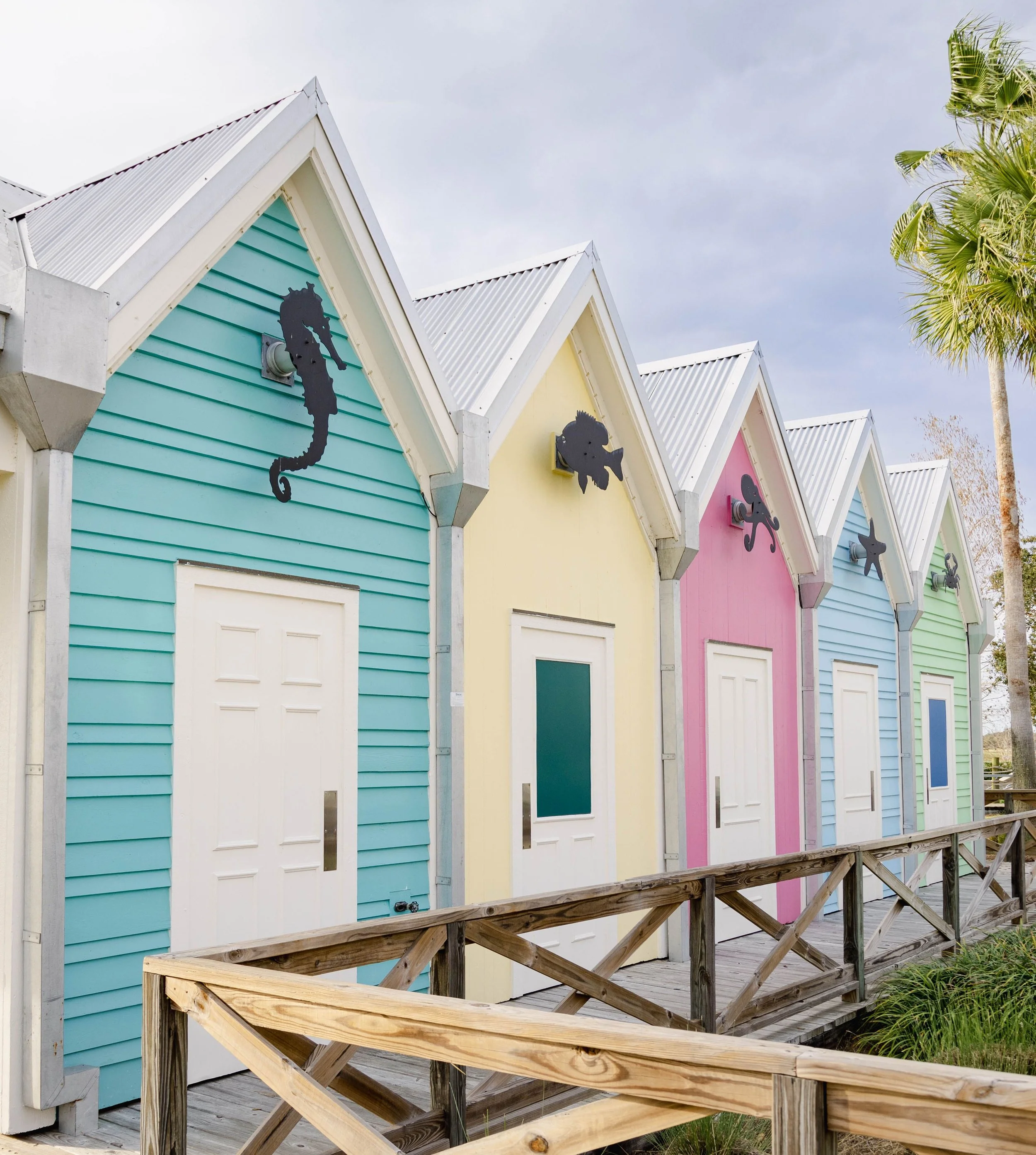 Colorful beach huts with sloped roofs, each painted a different pastel color such as blue, yellow, pink, and green, arranged in a row along a wooden walkway, with a partly cloudy sky and palm tree in the background.