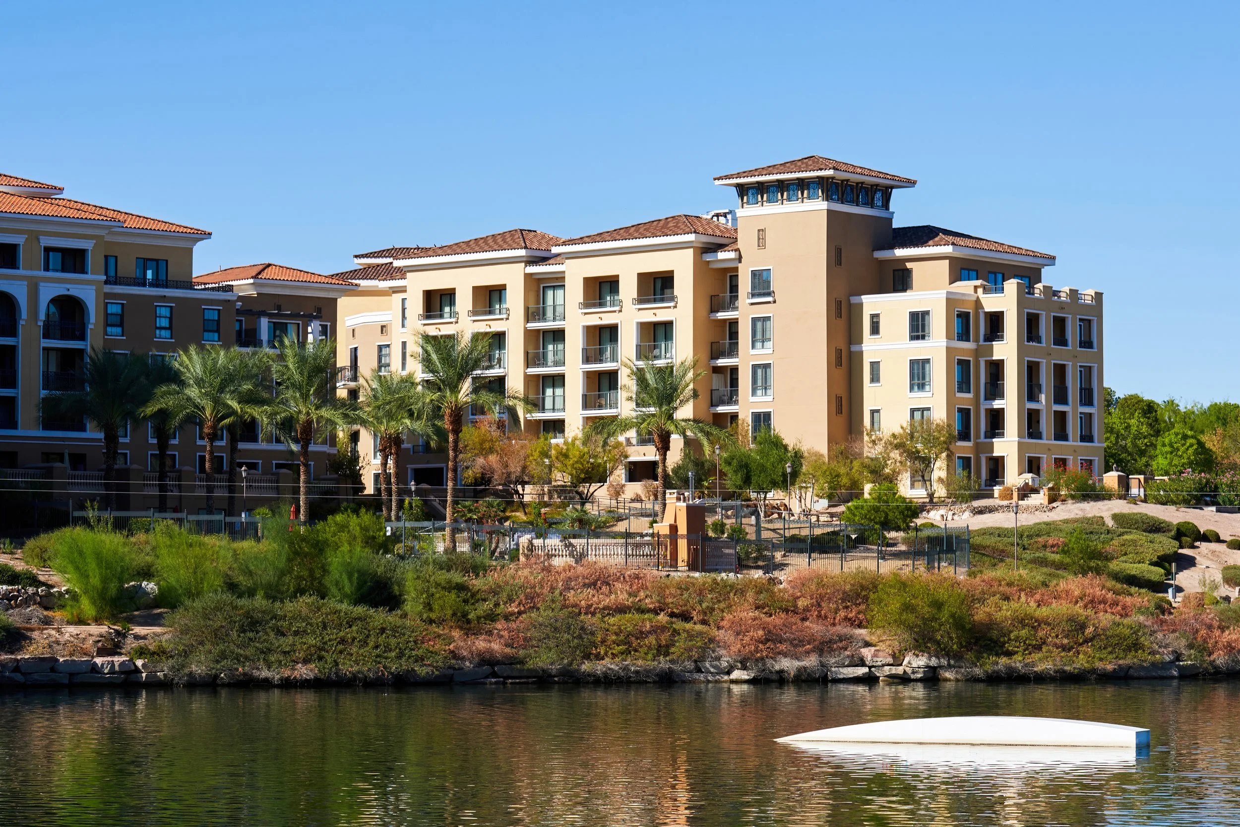A view of modern residential buildings with palm trees in front, overlooking a body of water under a clear blue sky.