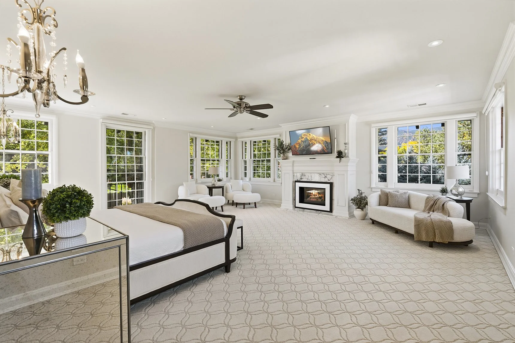 Bright living room with large windows, white walls, and a white carpeted floor. Features include a white sofa with a blanket, a glass-topped side table with a lamp and decorative plant, white armchairs by the windows, a fireplace with a mounted TV ab