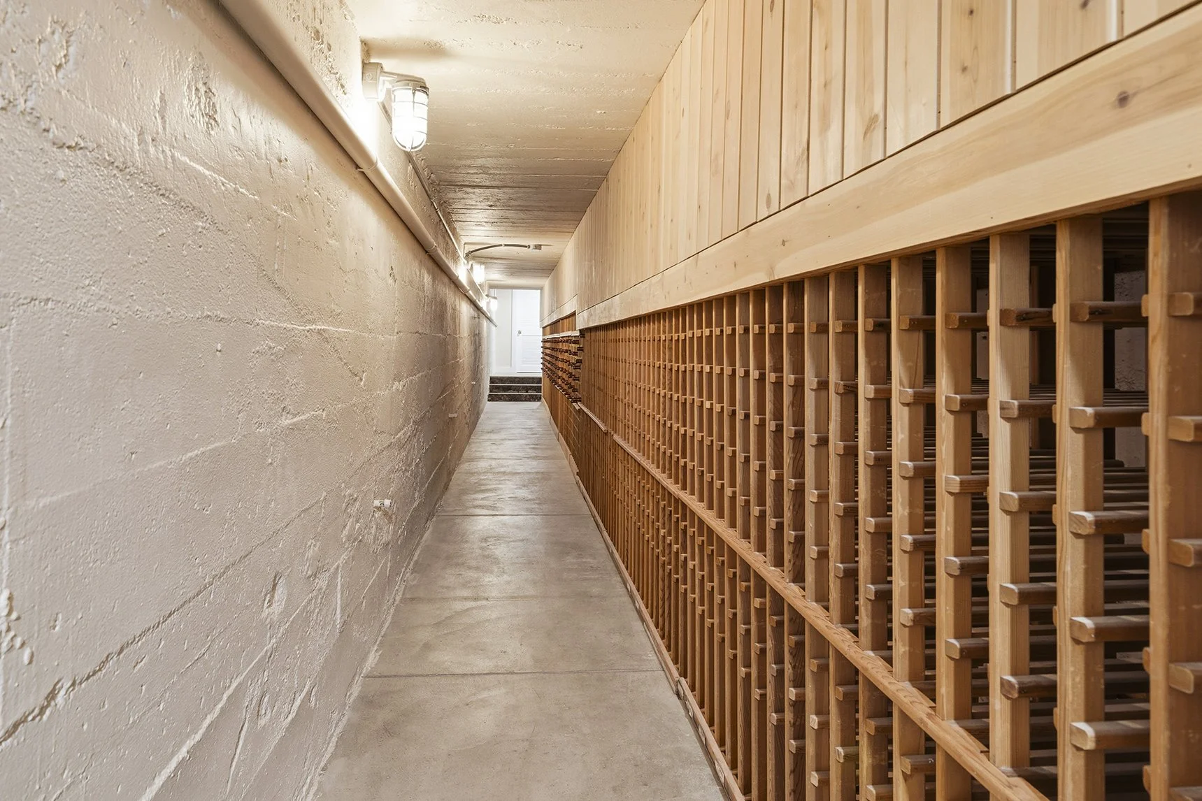 Narrow concrete basement hallway with wooden wine racks lining one wall and ceiling lights